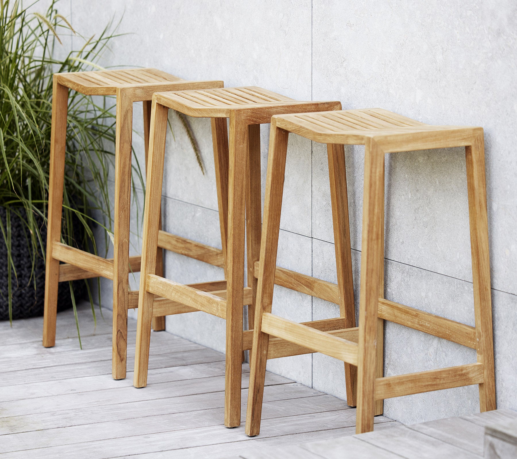 Tre træ flip-barstole med slidsede sæder, med et minimalistisk design og rene linjer, placeret ved siden af en dekorativ plante.