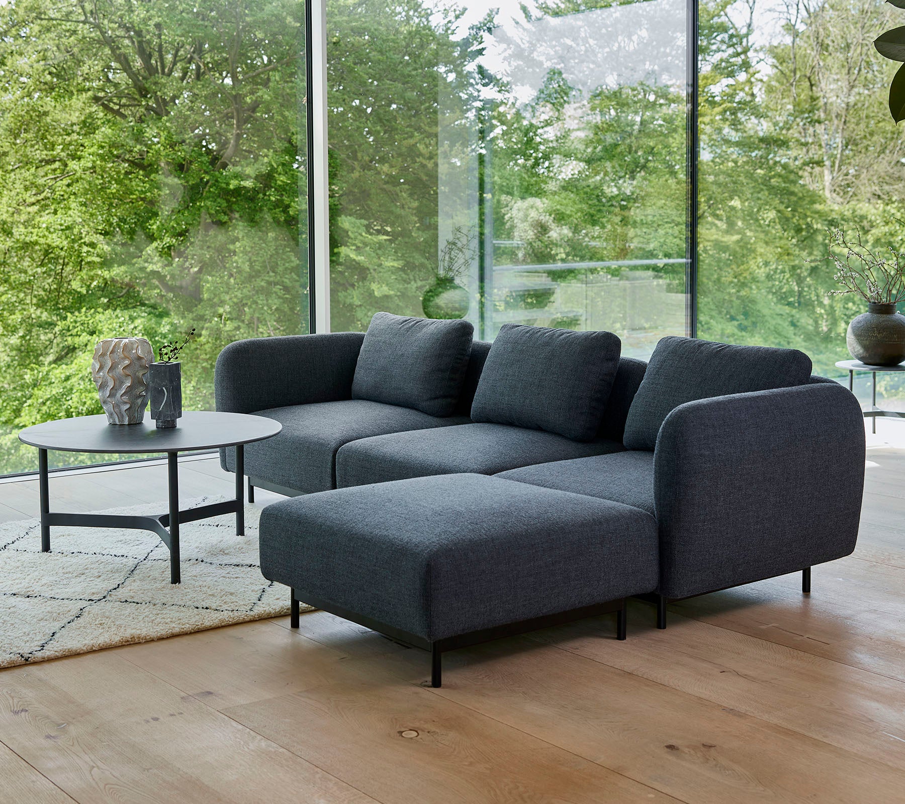 en moderne grå sektionel sofa med bløde puder, parret med et elegant rundt kaffe bord på et lyst tæppe, omgivet af grønne planter.