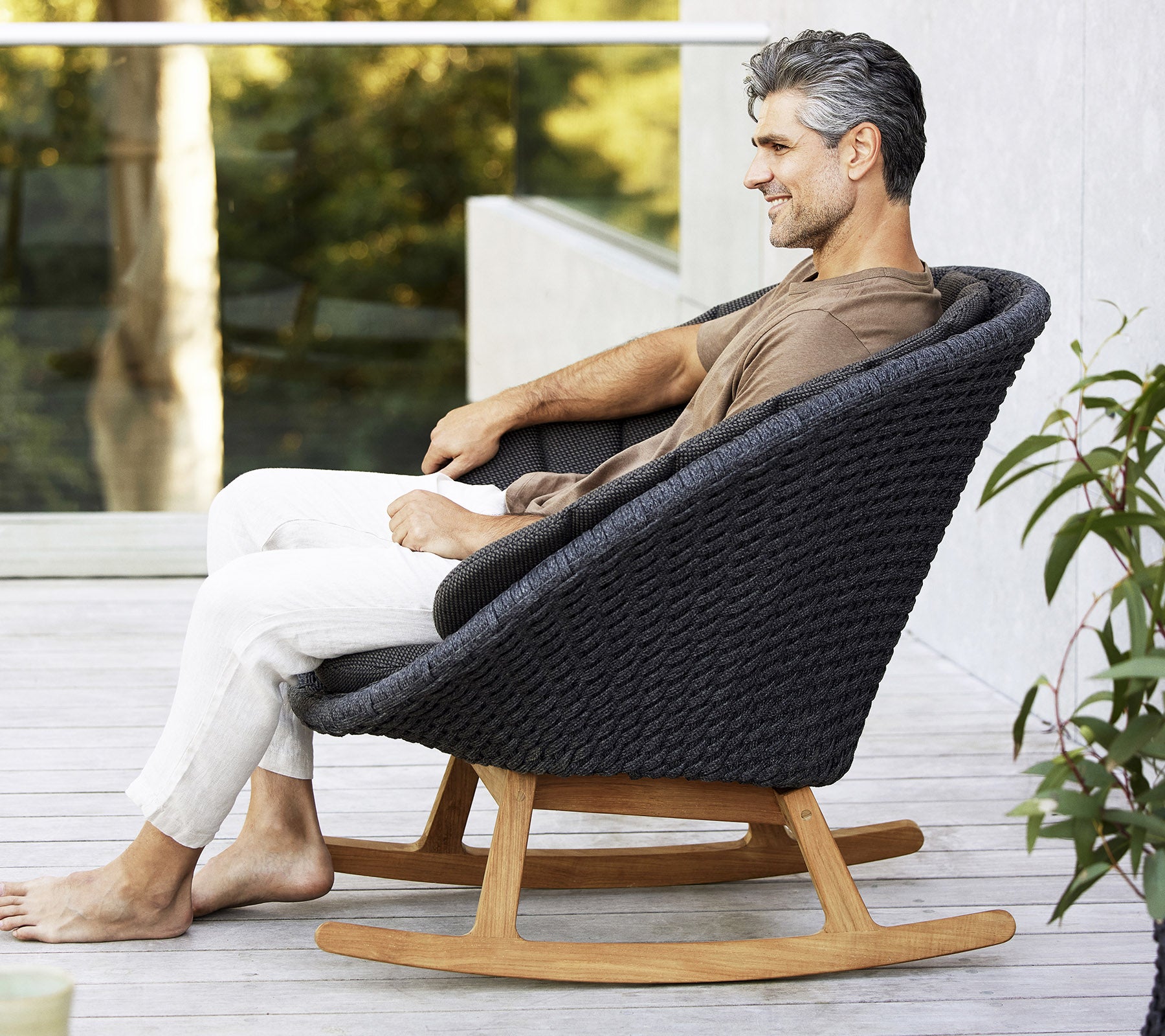 En mand sidder afslappet i en stilfuld, moderne gyngestol med et vævet design og en base af naturligt træ, omringet af grønne planter.