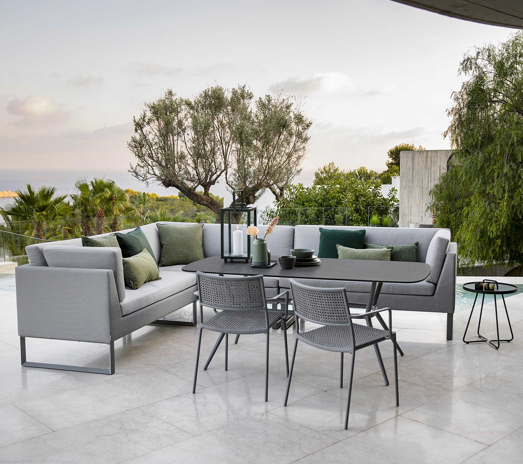 Moderne udendørs siddeområde med en stilfuld sektion sofa, et spisebord og metalstole omgivet af grønne planter.