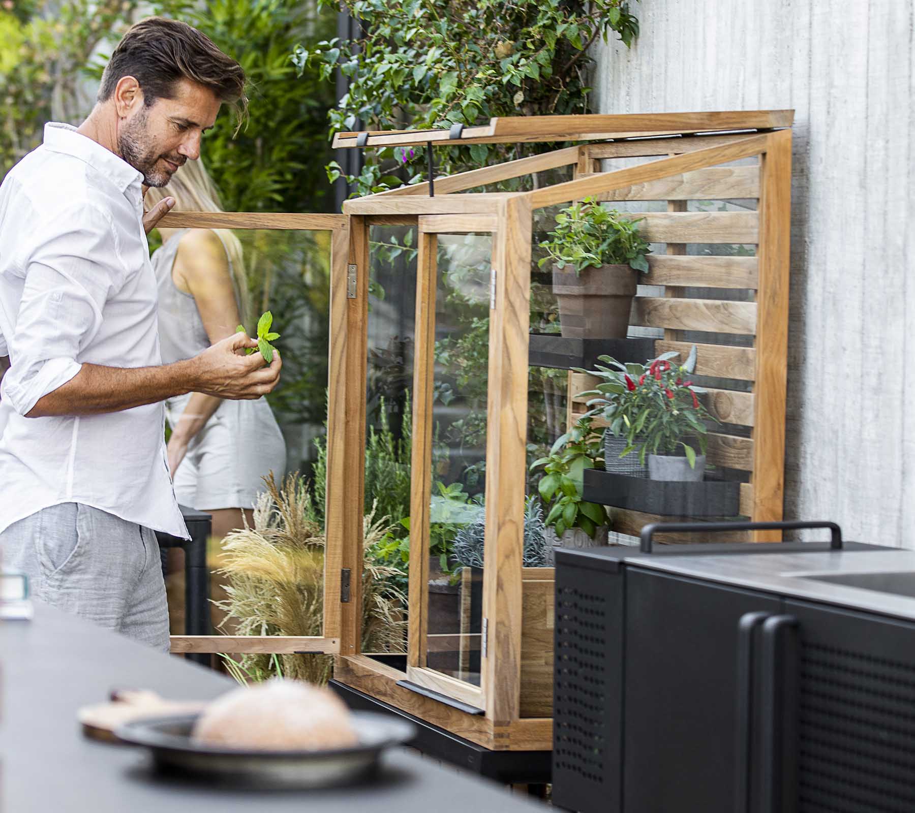 Mand, der passer planter i et glasdisplay, omgivet af grønne planter, med et moderne køkken i forgrunden.