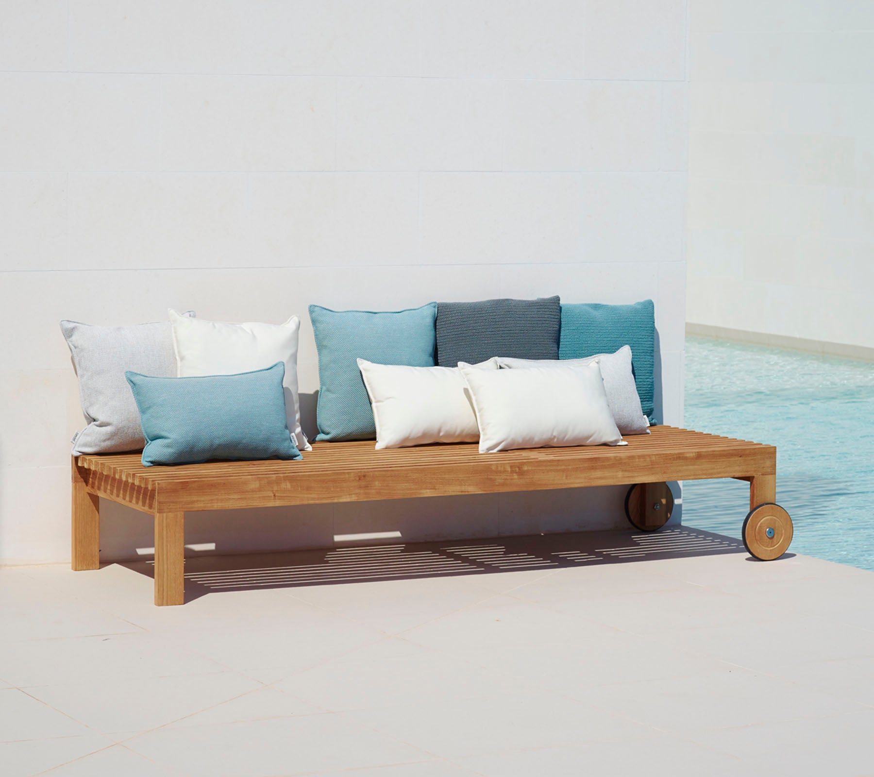 Træ loungesofa med enkelt design, udstyret med puder i nuancer af blå og hvid, placeret ved siden af en pool.