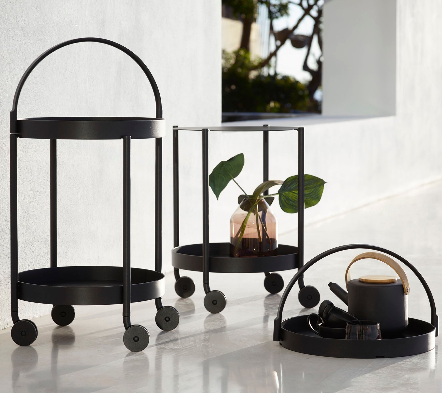 Tre sorte metalvogne med cirkulære rammer og hjul, der har en plante, dekorative genstande og en kop, arrangeret til fremvisning.