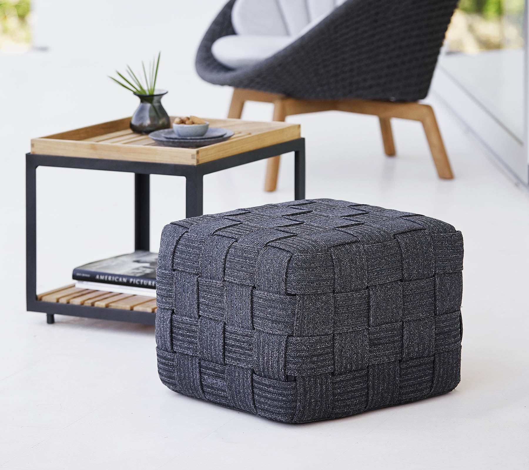 En struktureret grå vævet pouf placeret nær et træsidebord med et minimalistisk design, som indeholder små dekorationsgenstande.