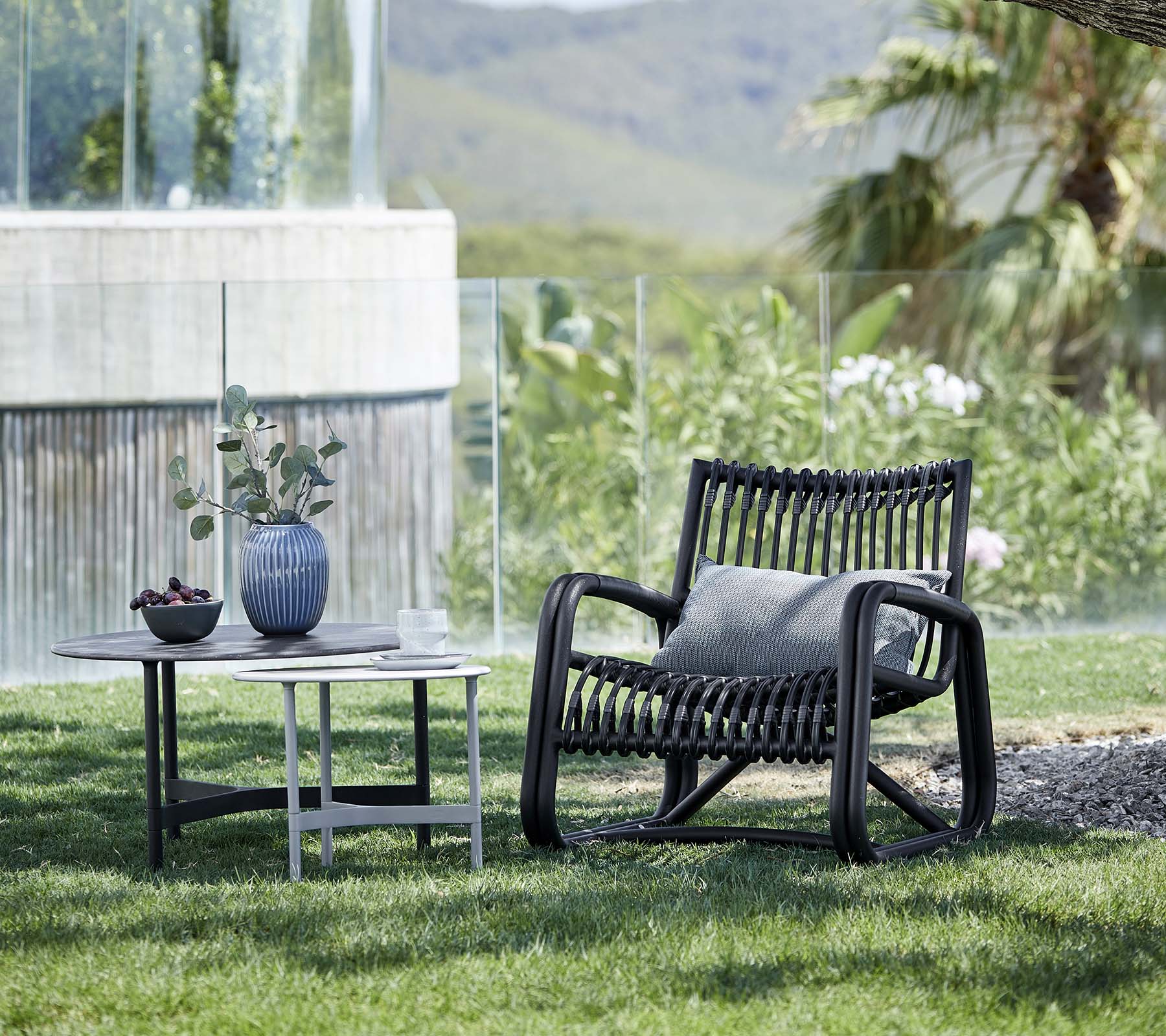 Samtidig loungestol med et mønstrede design, ledsaget af et sidebord med en vase og en potteplante, placeret i et naturligt landskab.