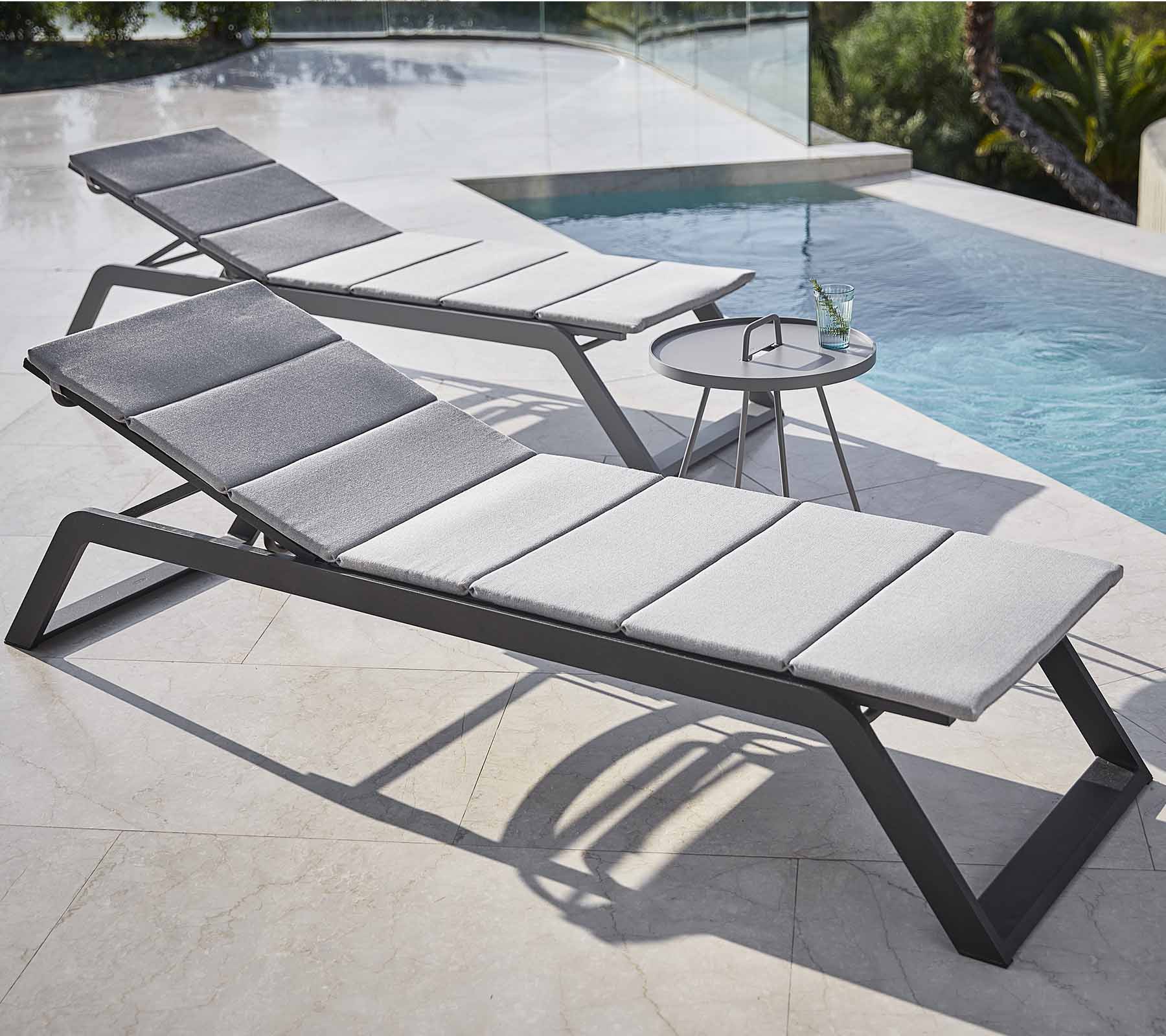 To stilrene loungestole med grå puder er placeret ved en pool, ledsaget af et lille sidetable med en drink.