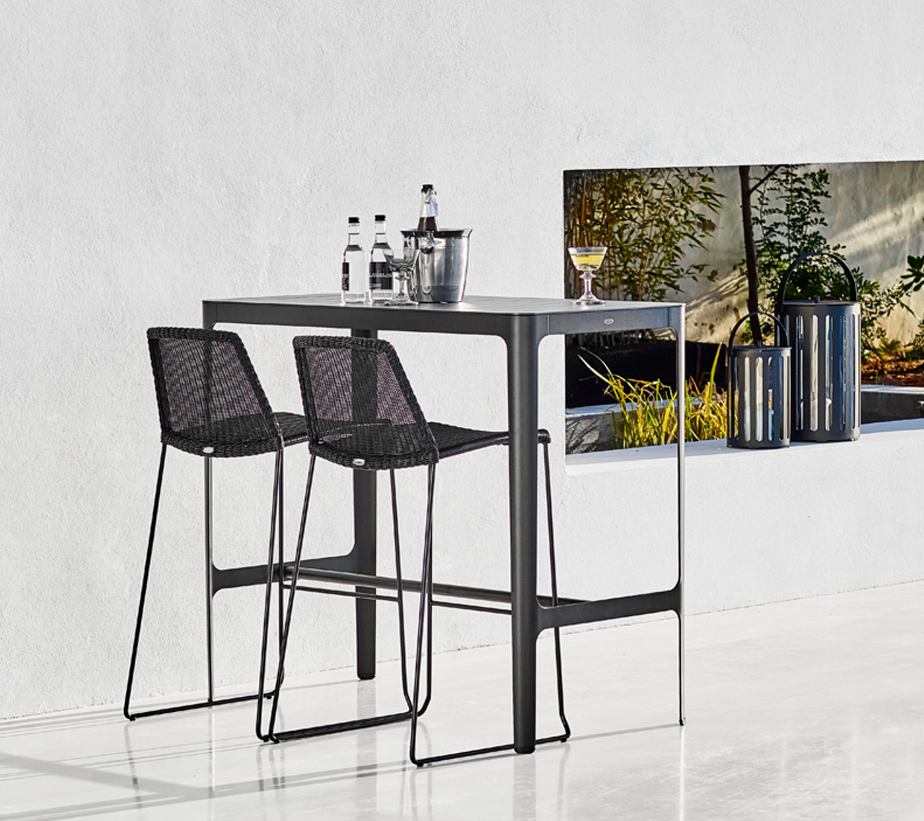Elegant bardisk med to sorte vævede barstole, ledsaget af glasvarer og en stilfuld spand, sat mod en minimalistisk baggrund.