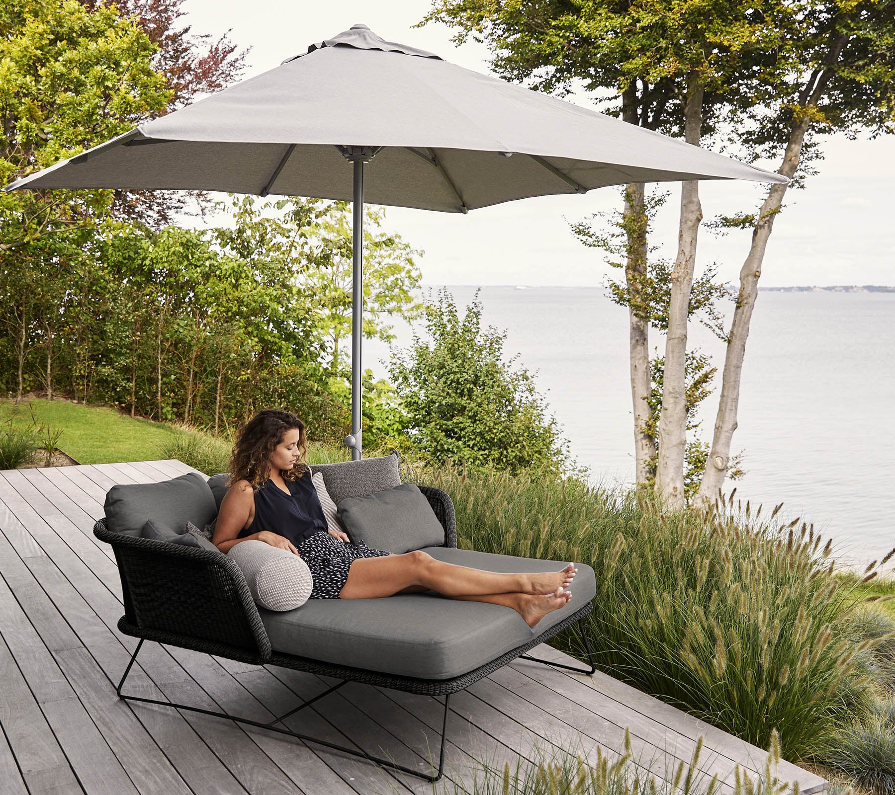 En kvinde slapper af i en moderne liggestol med en stor parasol, omgivet af grønt og med udsigt til vandet.