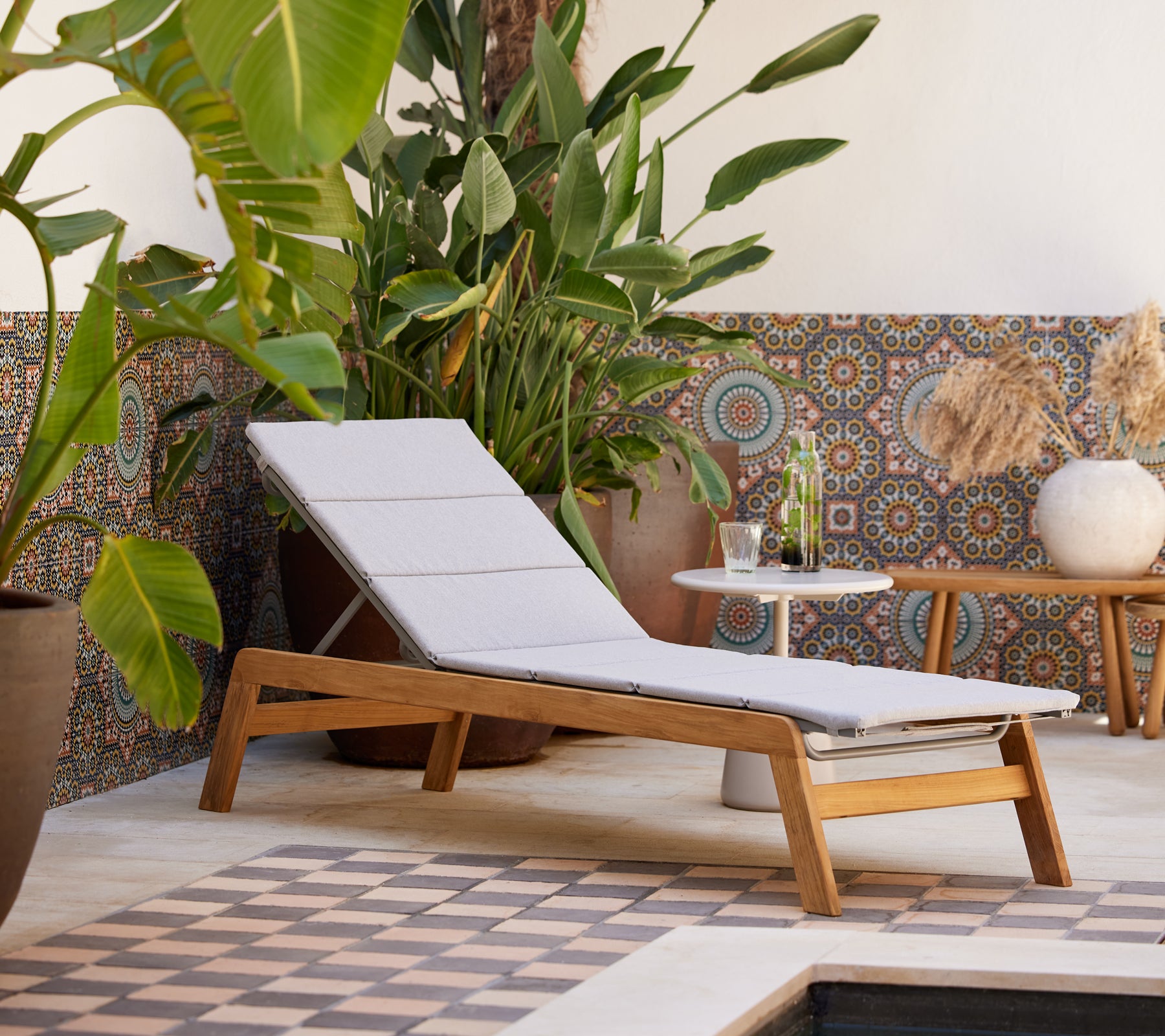 En moderne lounge stol med en lys pude, parret med et sidebord, omgivet af frodig grøn vegetation og dekoreret flisebaggrund.