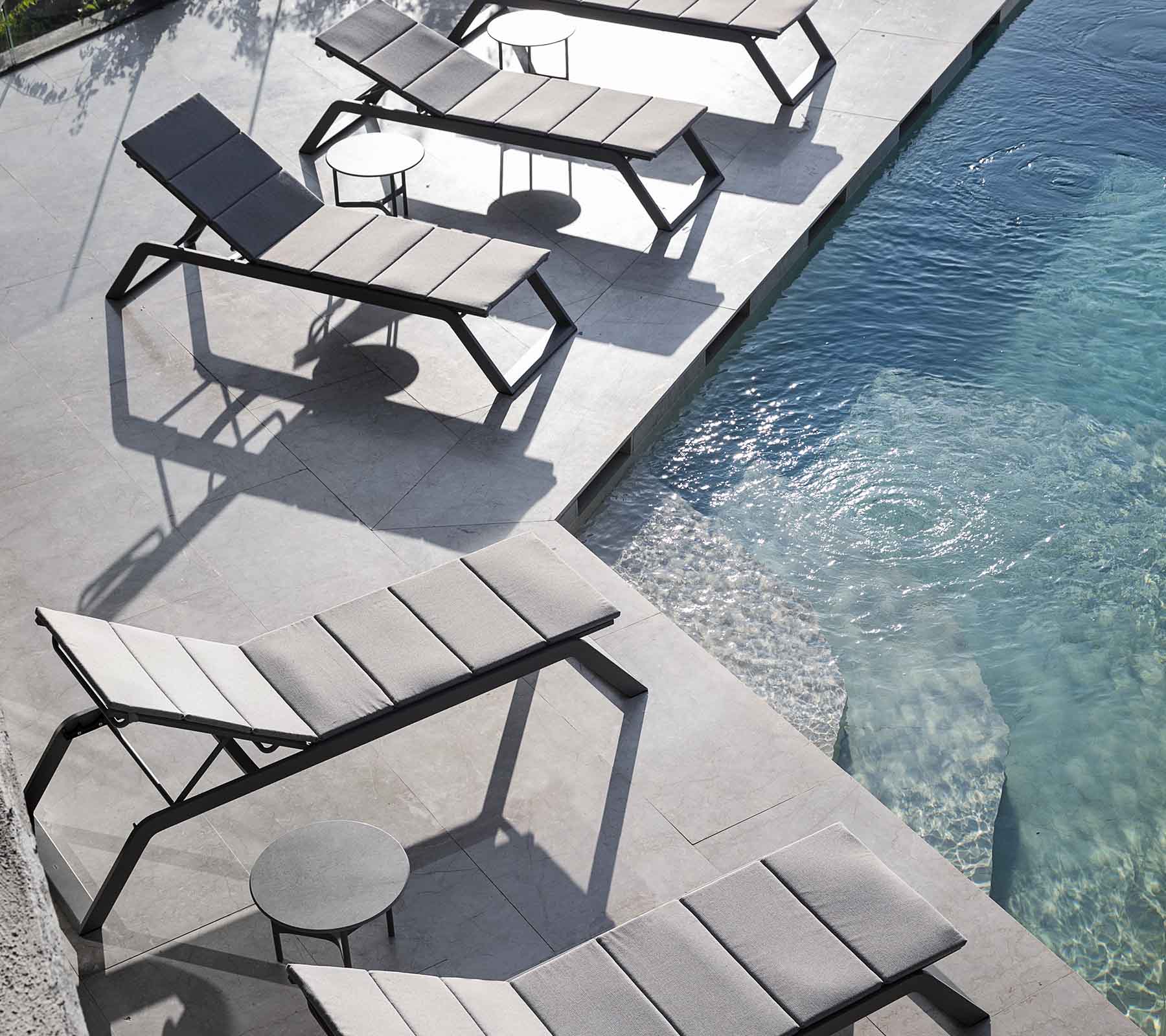 En række moderne lounge-stole med et lille sidebord ved siden af en swimmingpool, der kaster skygger på fortovet.