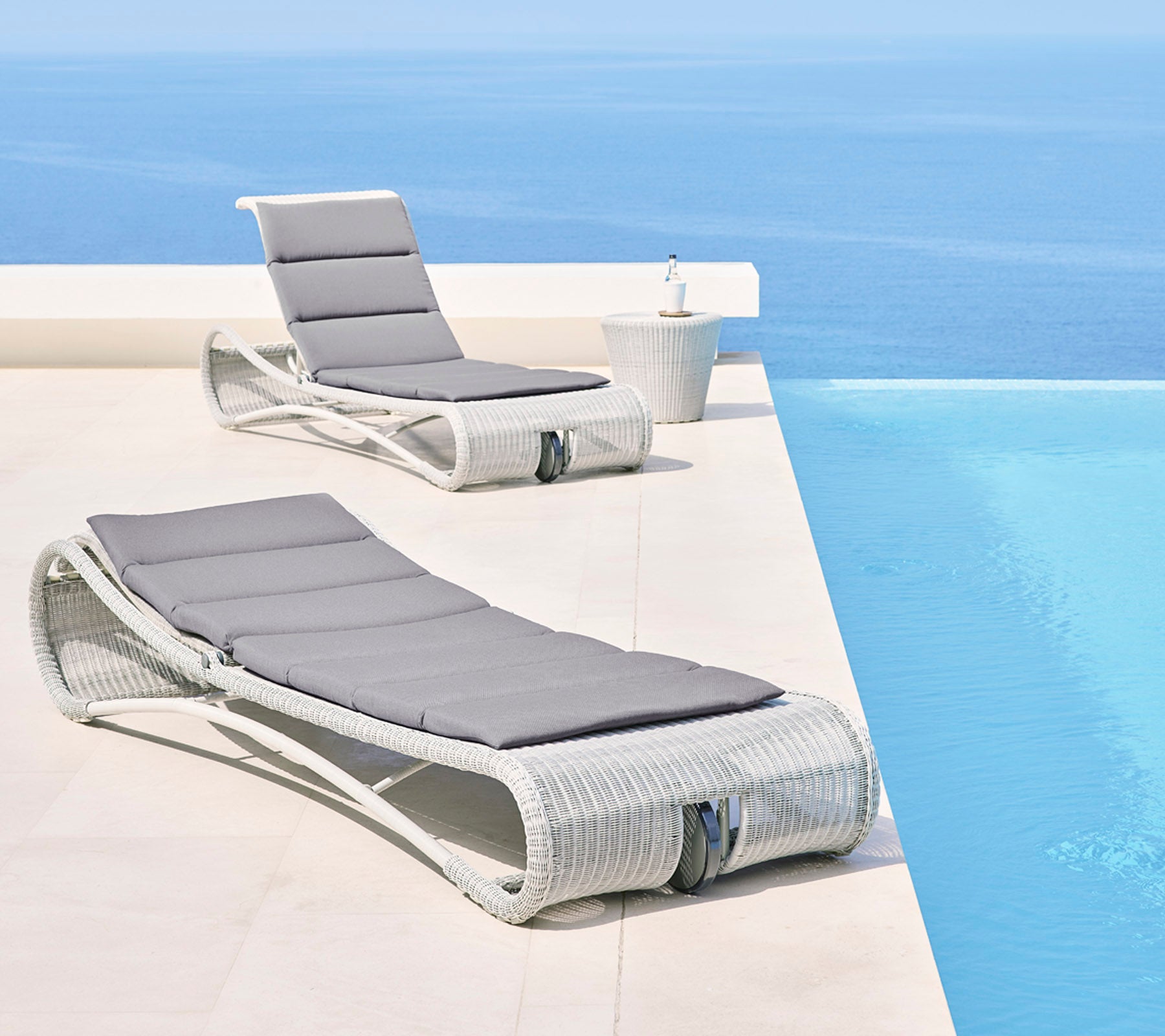 To stilfulde loungestole med grå puder ved en fredfyldt blå infinity-pool, der skaber et afslappende udendørsområde.