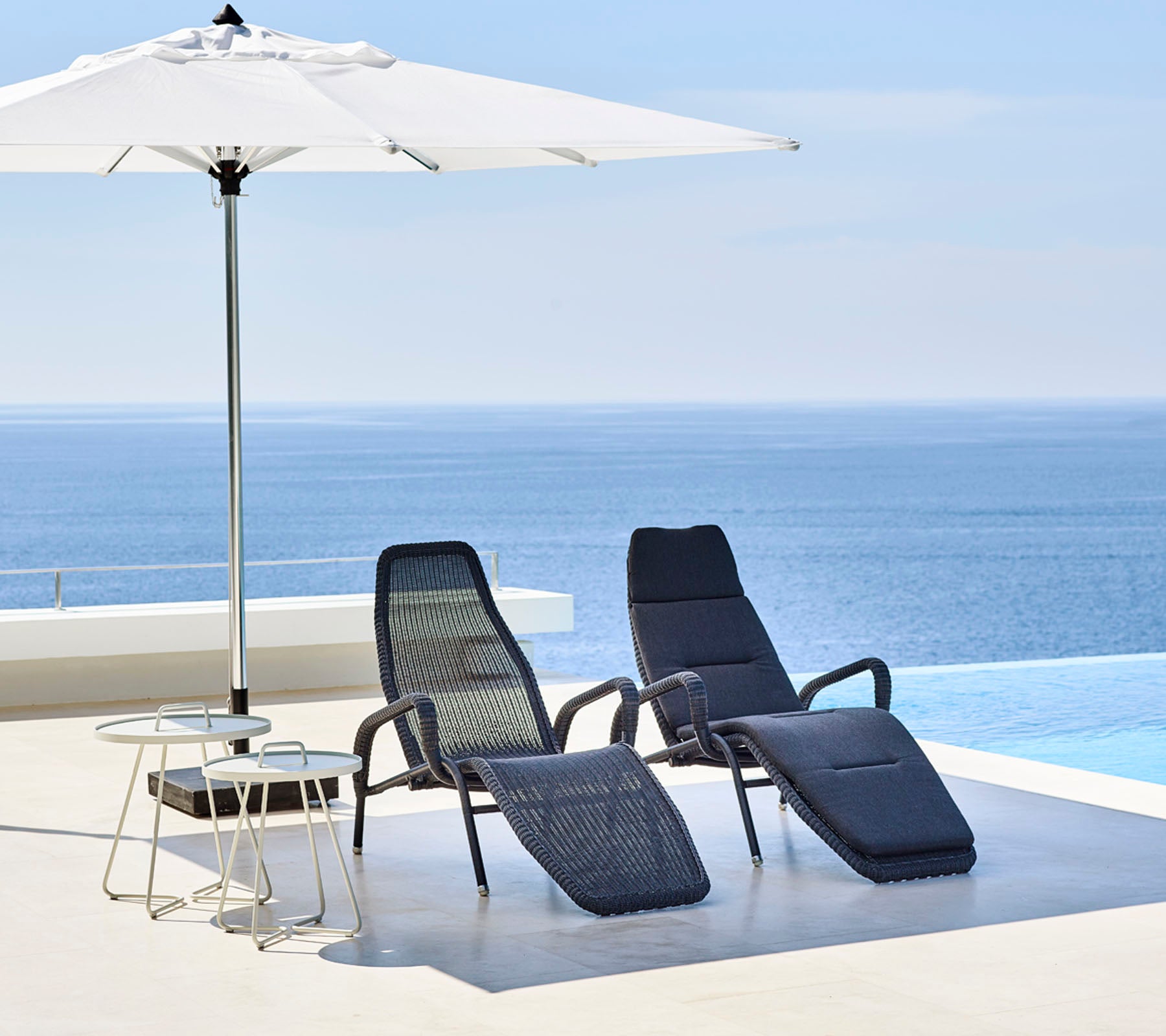 To stilfulde loungestole og et lille bord står under en stor parasol med et roligt blåt hav i baggrunden.