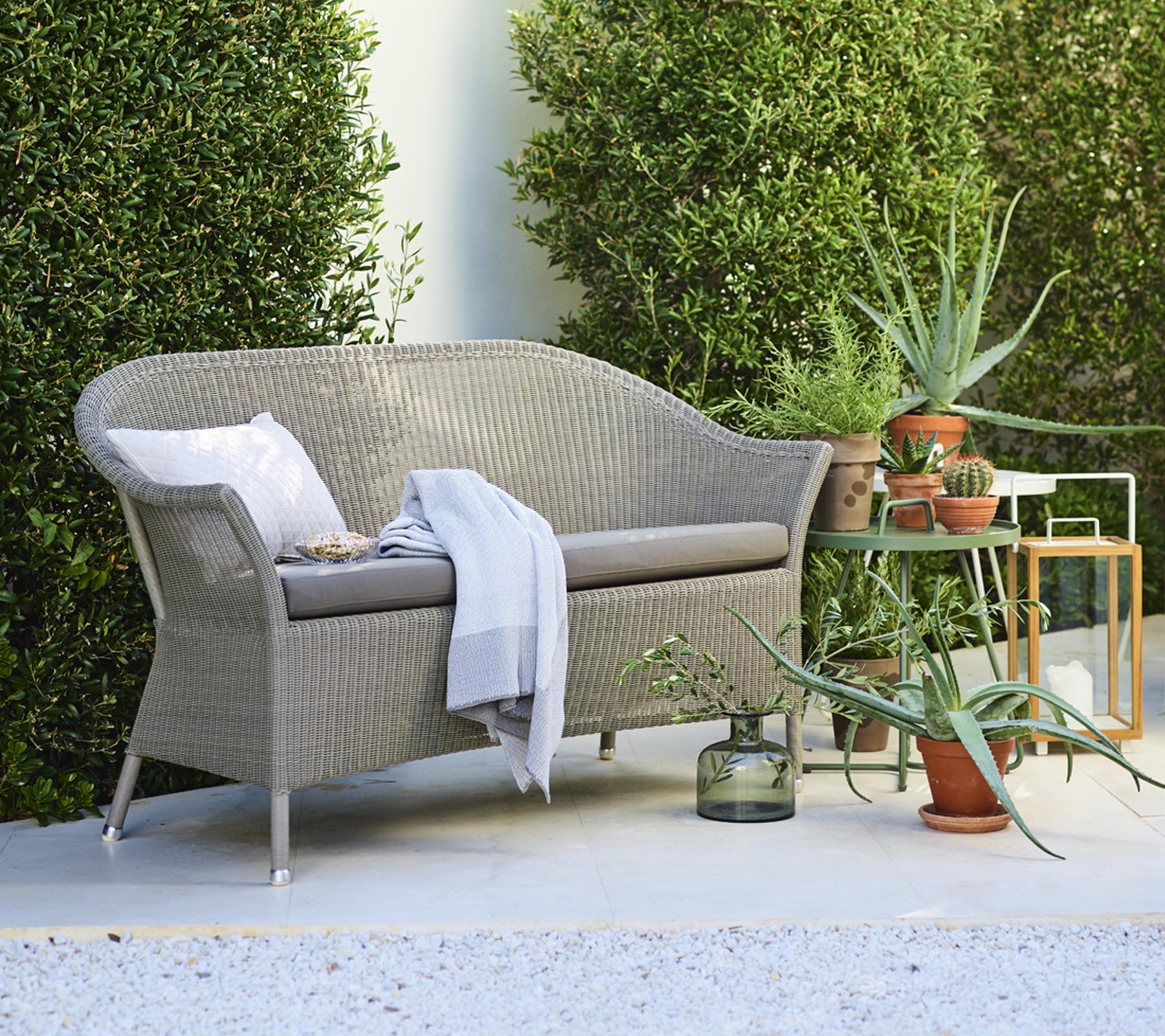 En hyggelig loveseat med dekorative puder, ledsaget af potteplanter og et sidebord, omgivet af grønt.