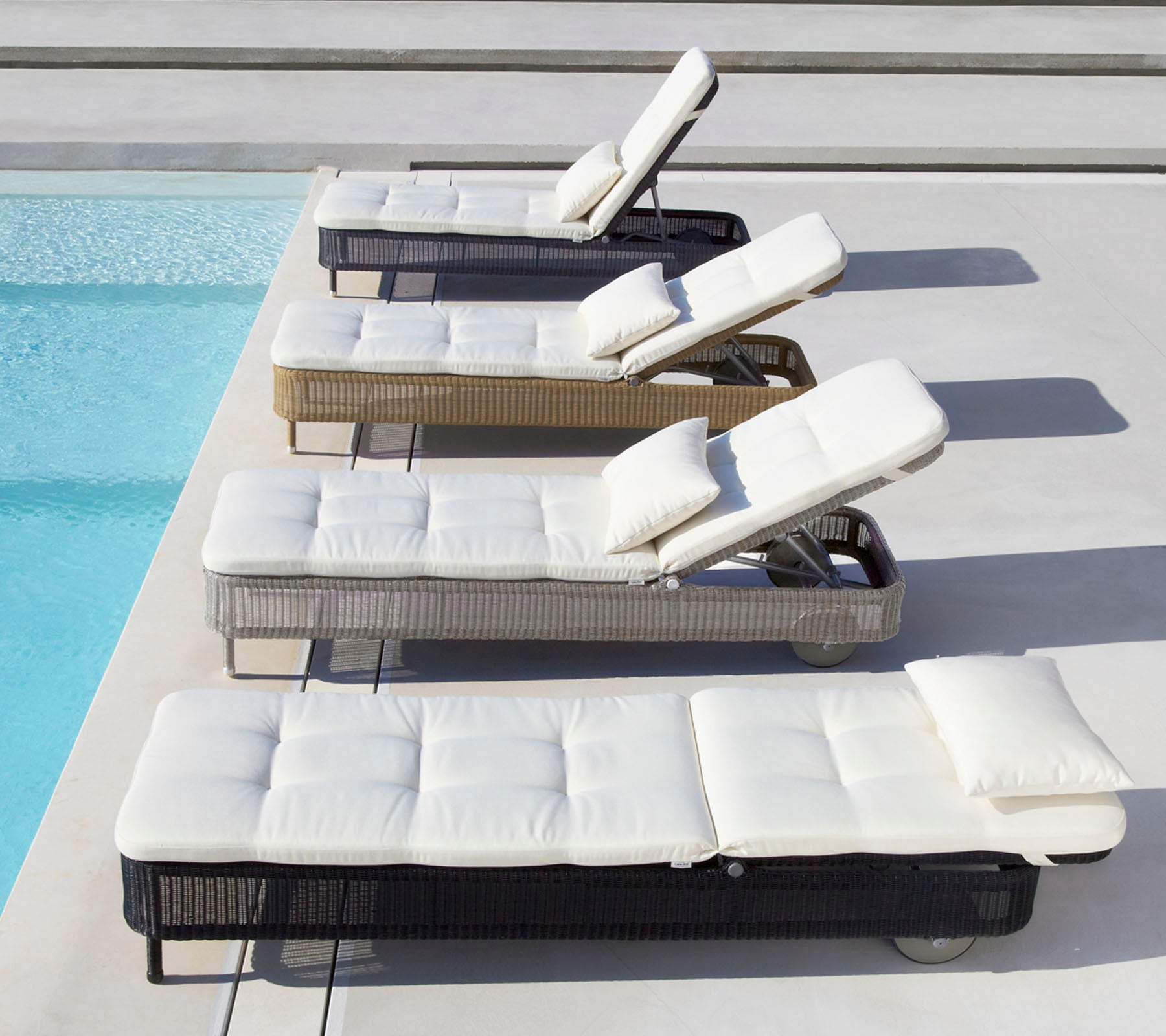 Fire liggestole i forskellige farver med hvide puder, placeret ved siden af en pool, hver med justerbar ryg.