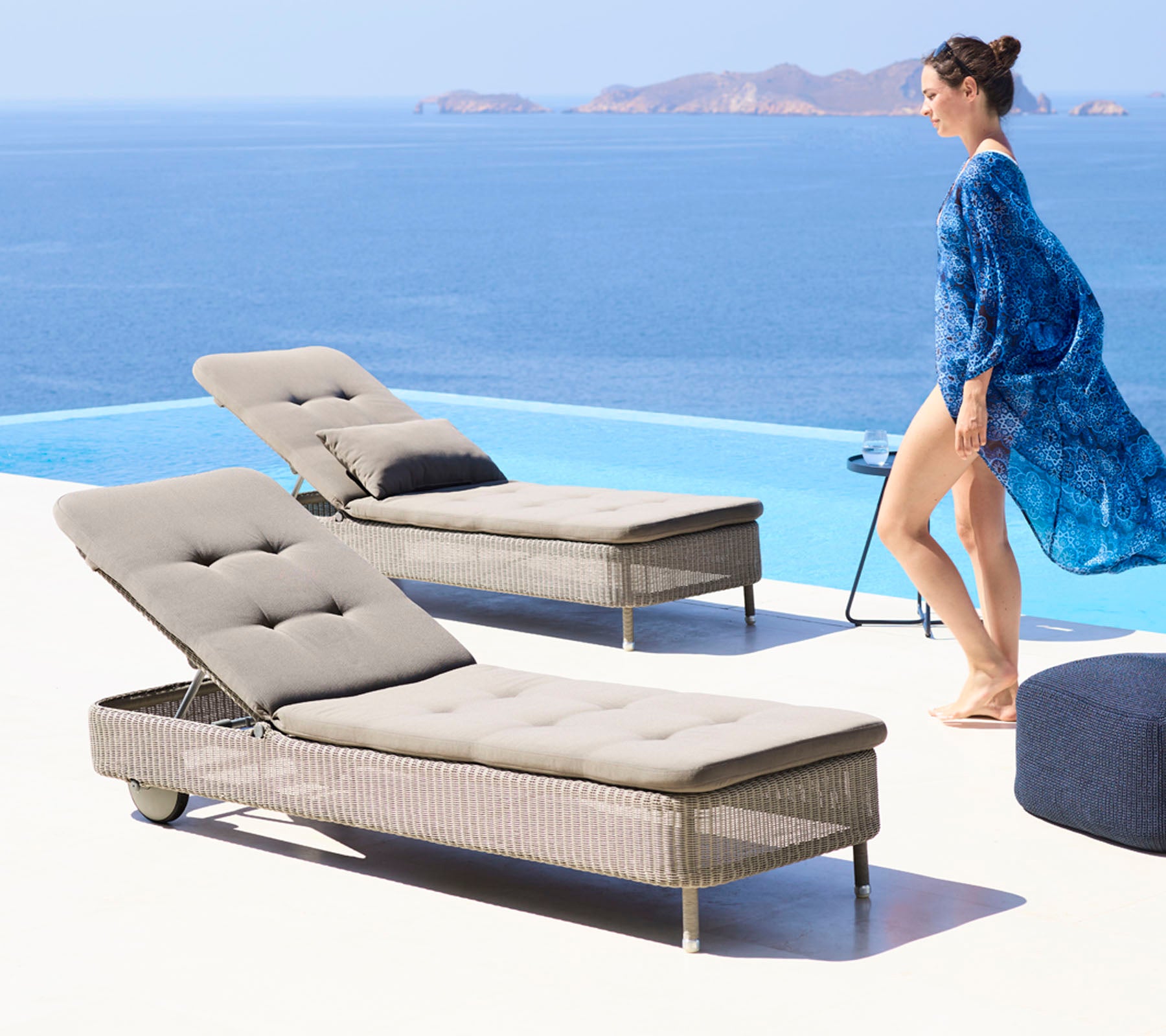 En kvinde i en flydende blå cover-up går ved siden af to stilfulde lounge stole ved en pool med udsigt til et roligt hav.