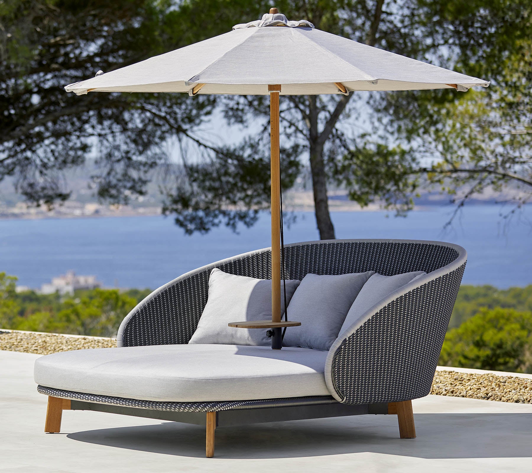 Stilfuld udendørs loungestol med komfortable puder og en stor parasol til skygge, sat mod en naturskøn baggrund.