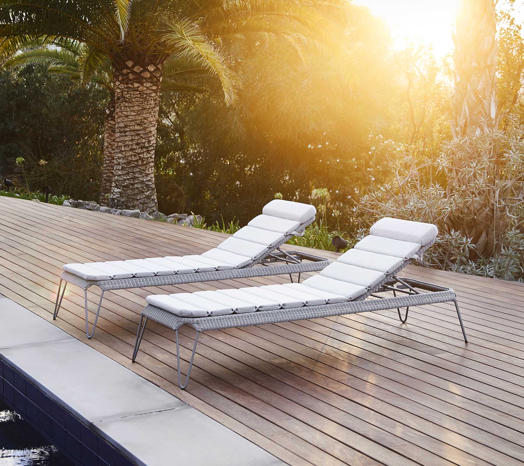 To stilfulde loungestole med hvide puder, placeret ved siden af en pool og omgivet af frodig grøn vegetation, der nyder den varme sollys.