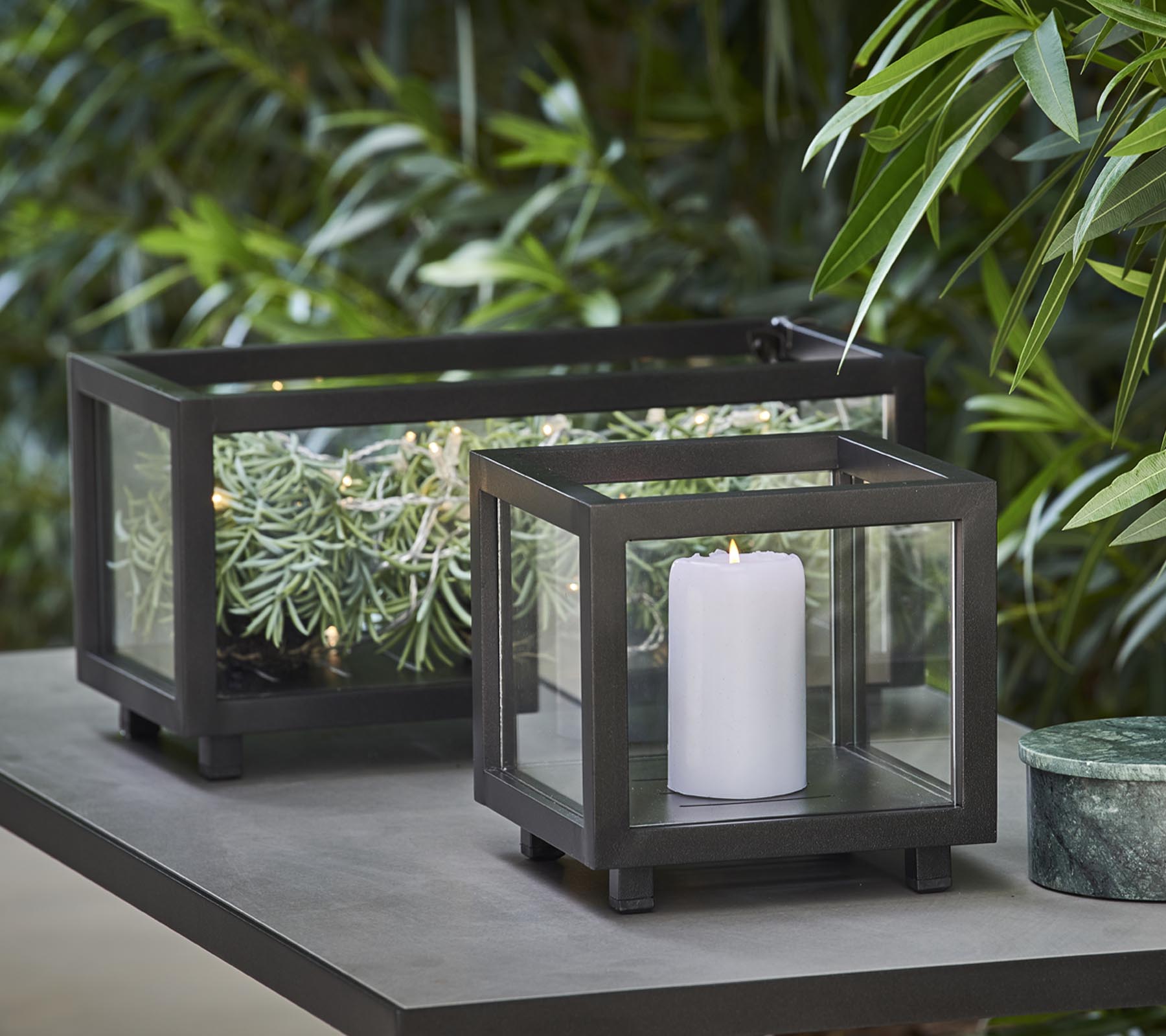 To stilfulde lanterner med glaspaneler, den ene med et hvidt lys og den anden med grønne planter, placeret på et bord midt i frodig løv.