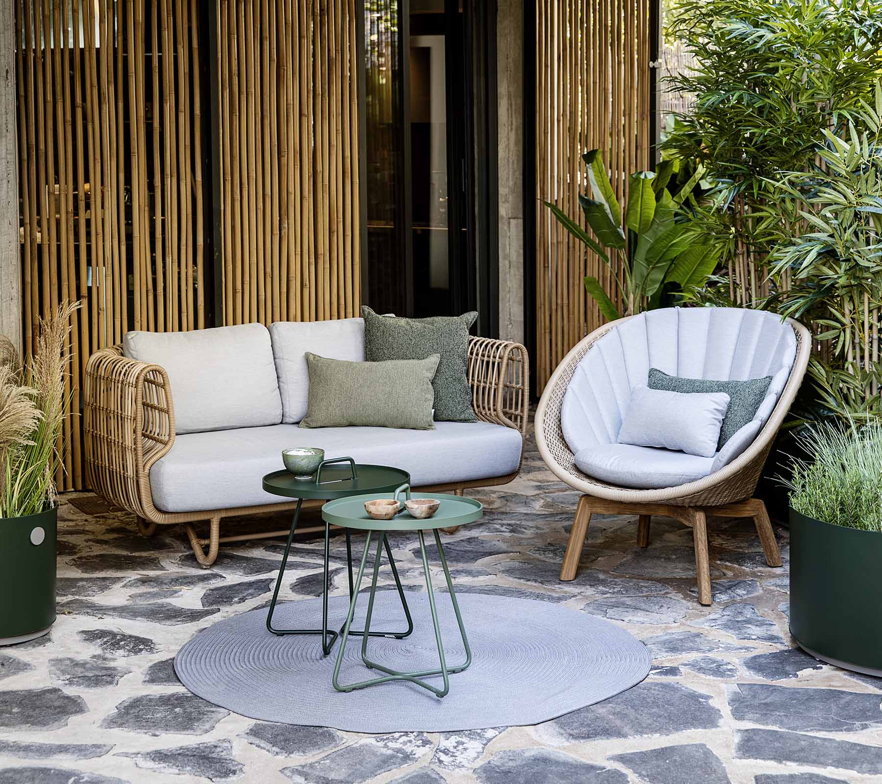 Et hyggeligt siddearrangement med en rattansofa og en stilfuld stol, ledsaget af et lille bord og dekorative planter.
