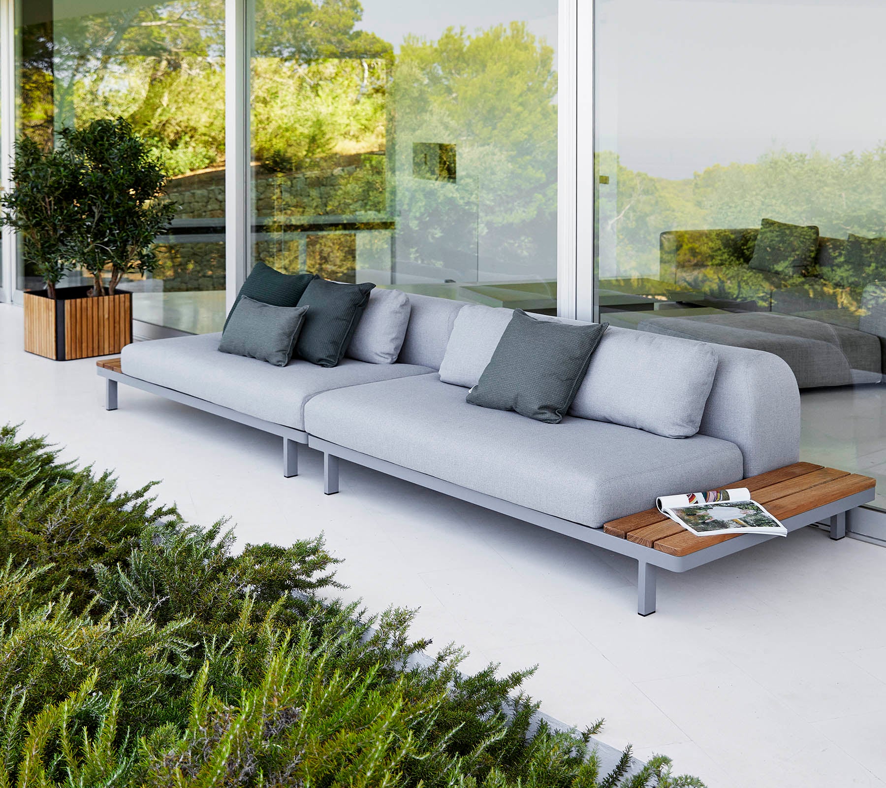 Moderne sofa med lysegrå puder og trædetaljer, ledsaget af dekorative puder og omringet af grønne planter.