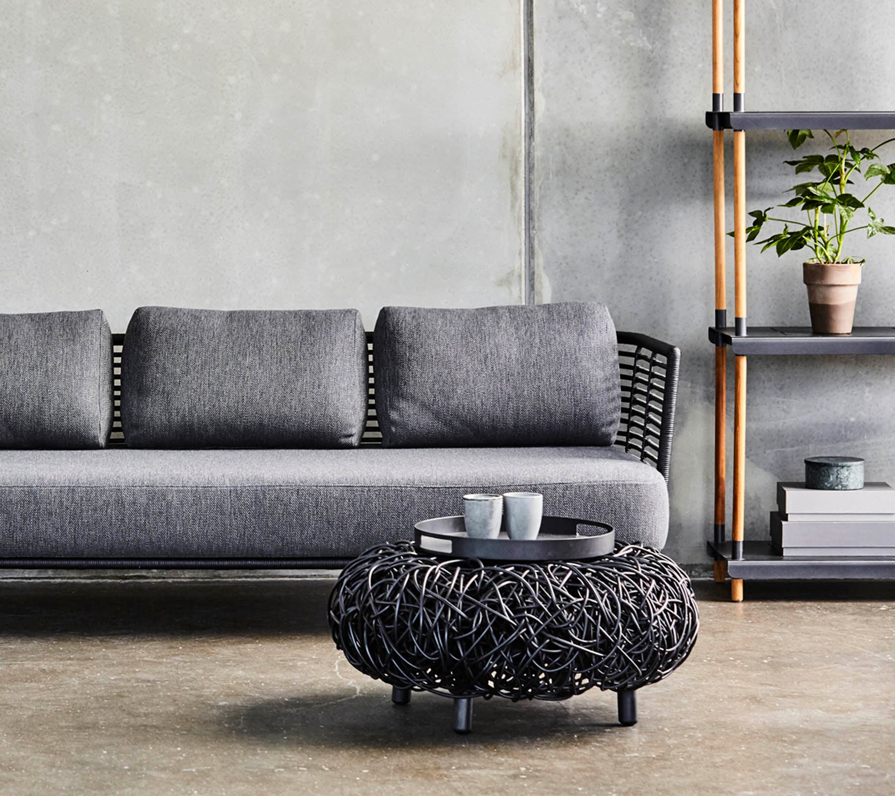 En moderne grå sofa med puder, parret med et cirkulært sort vævet kaffebord og en minimalistisk hylde, der viser planter og dekoration.