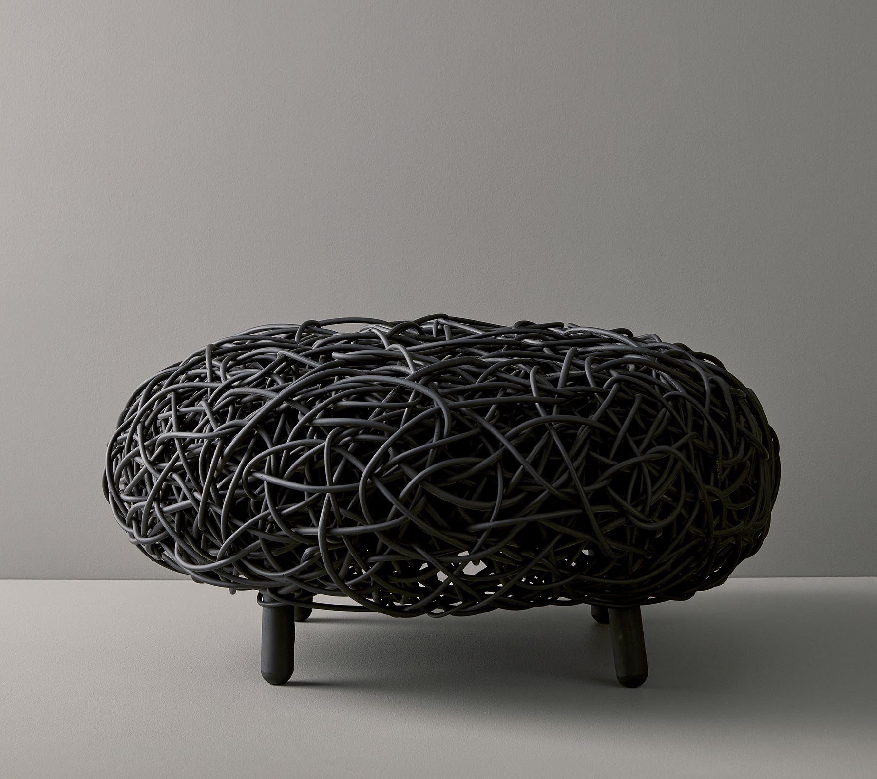 Sort vævet ottoman med en organisk, rund form og indviklet design, hvilende på tre slanke ben mod en neutral baggrund.