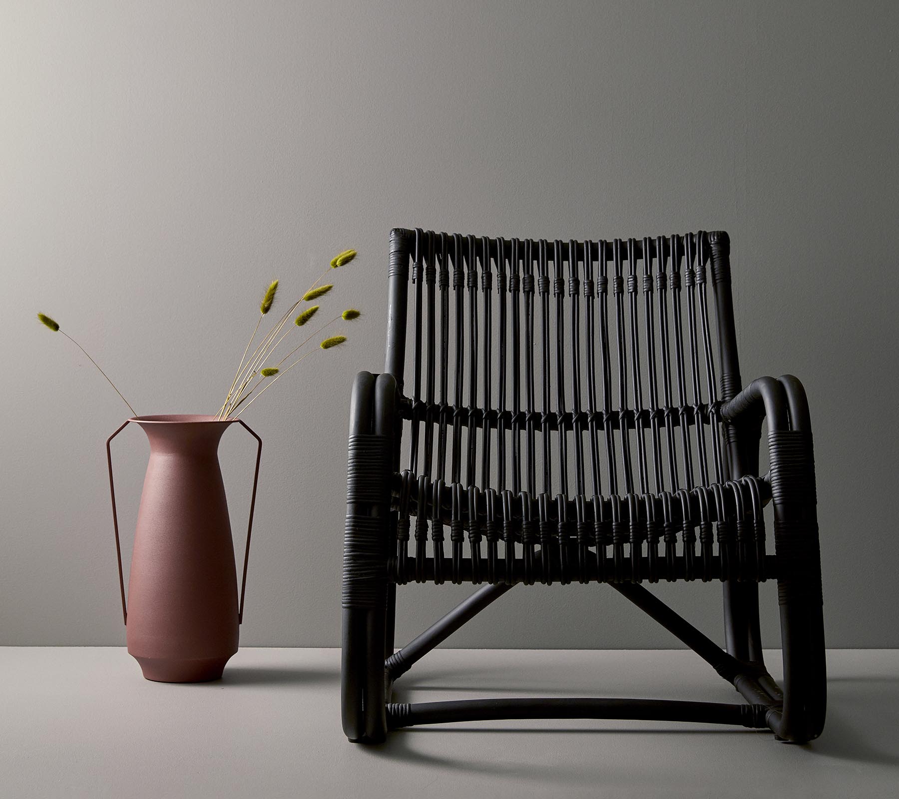 Stilfuld sort vævet stol ved siden af en lyserød vase med grønne stængler, sat op mod en minimalistisk grå baggrund.