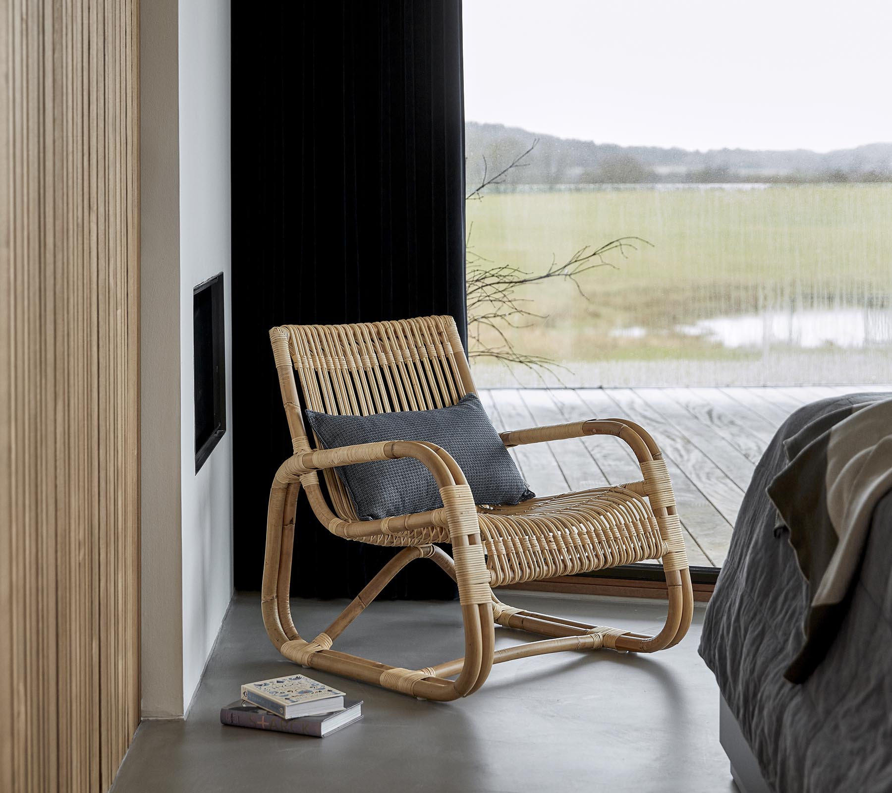 En stilfuld gyngestol lavet af naturmaterialer, placeret ved siden af en seng og en bog, med en naturskøn udsigt synlig gennem store vinduer.