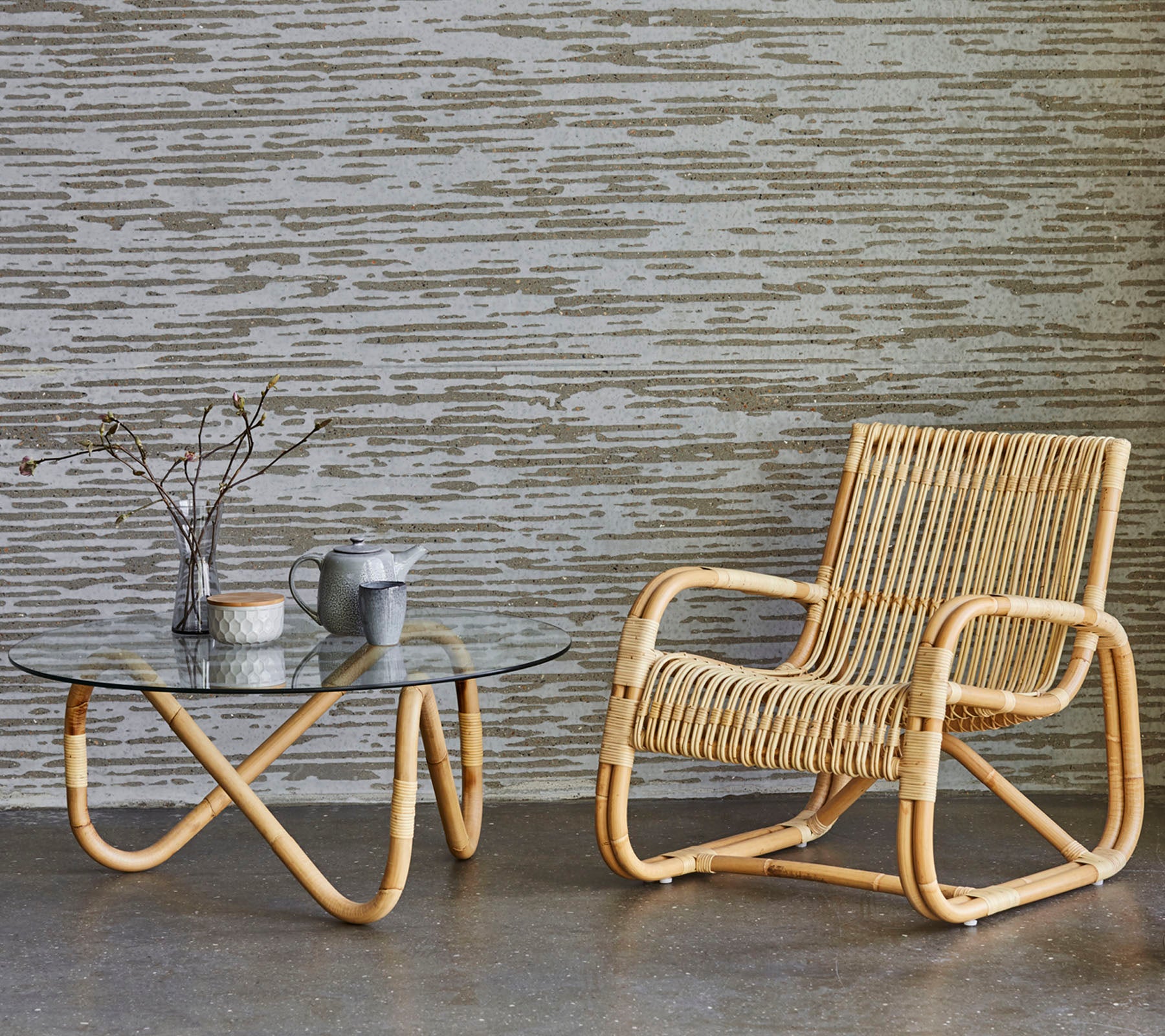 Rattanstol og glasbord med dekorative genstande, sat op mod en tekstureret væg. En minimalistisk og moderne indretningsstil.