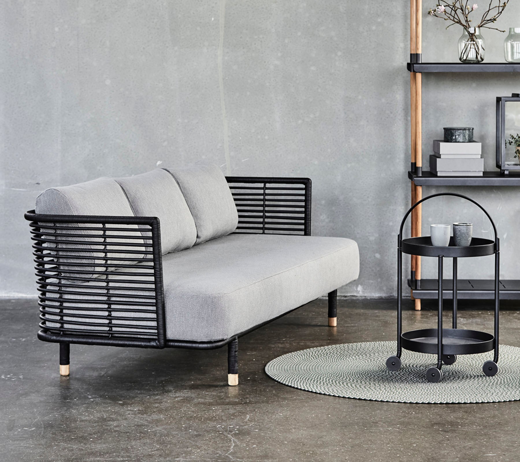 Slank grå sofa med sorte metal accenter og puder, parret med et rundt siddebord på et tekstureret tæppe mod en neutral baggrund.