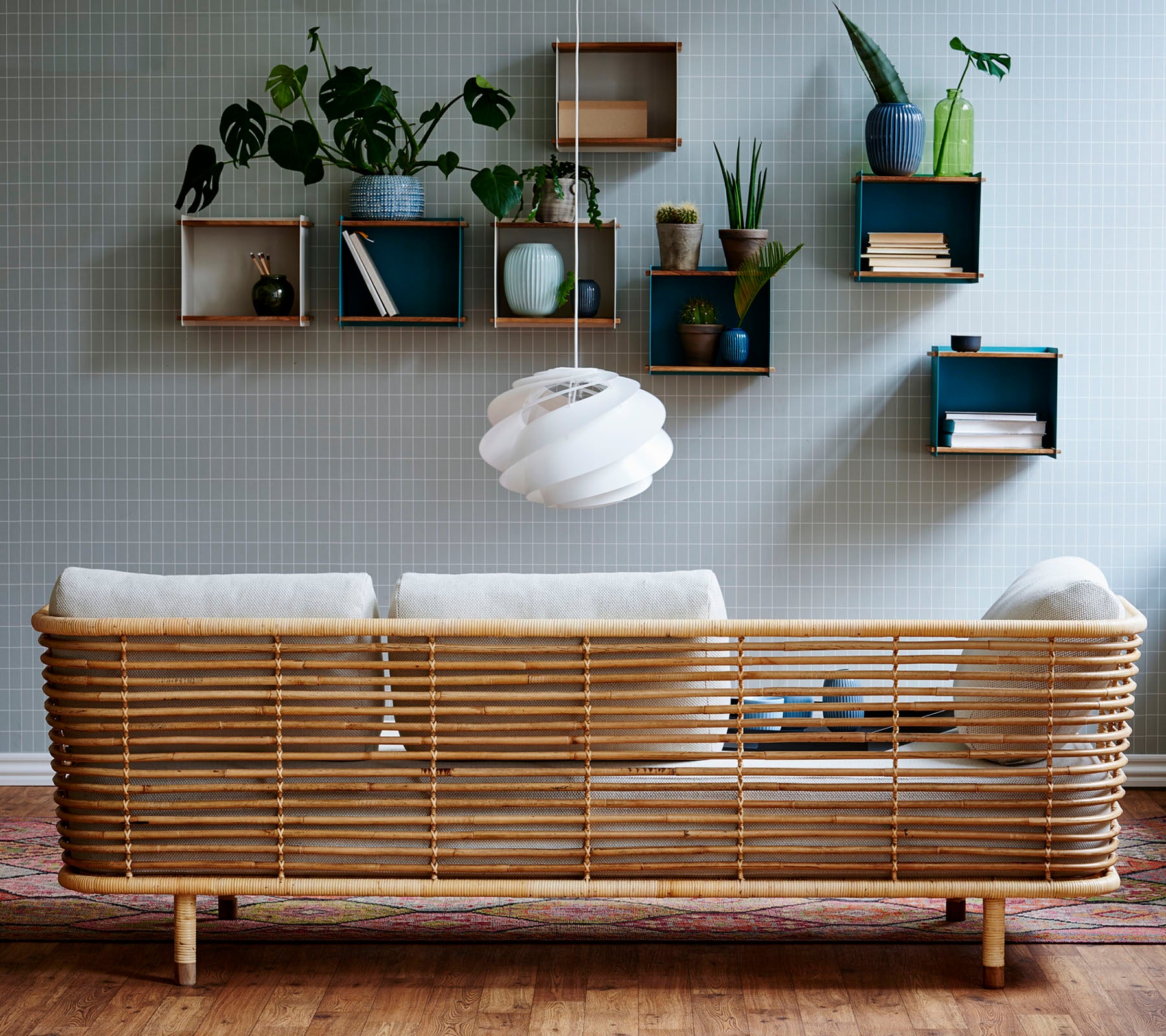 En stilfuld rattan-sofa med lyse puder mod en struktureret væg, suppleret af væghængte hylder, der viser planter og dekorative genstande.