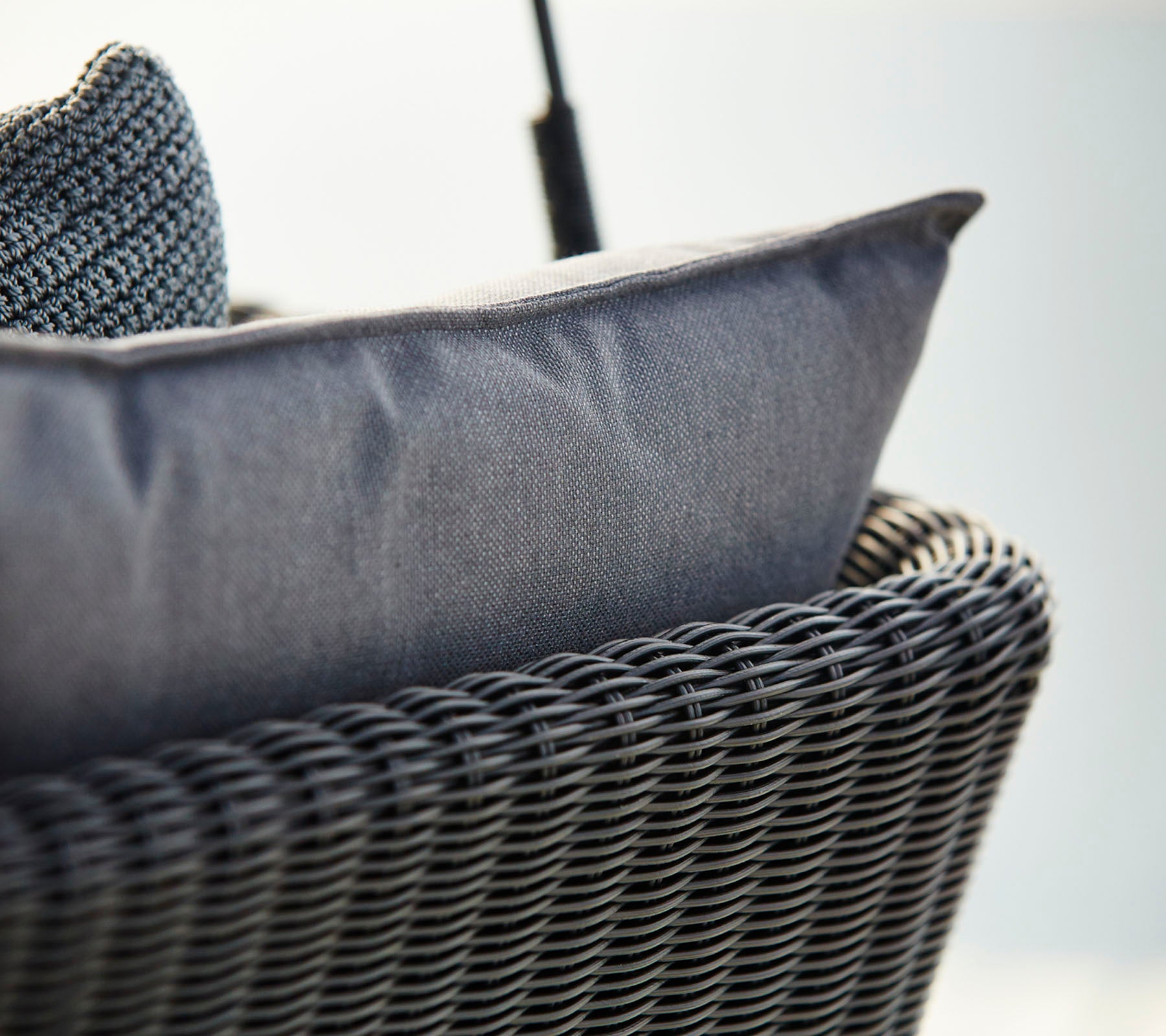 Close-up af en vævet kurvestol med en tekstureret mørkegrå pude, der understreger komfort og moderne design.