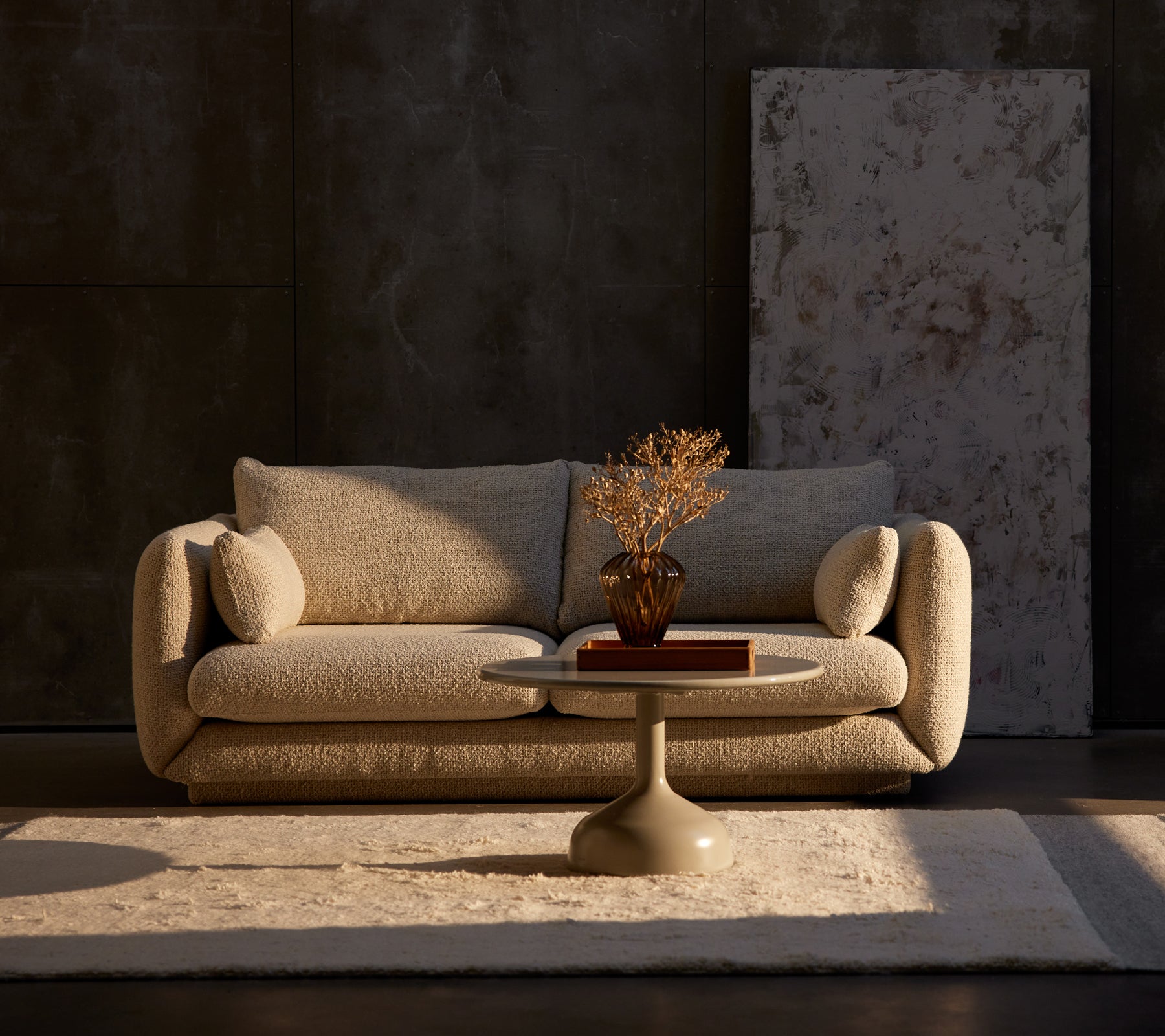 Bloom 2-personers sofa i sandfarvet flair med et Glaze kaffebord og en dekorativ plante på et tæppe, sat mod en struktureret mørk væg.