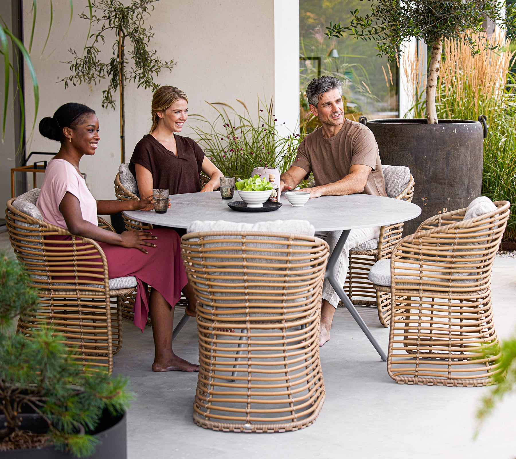 Fire personer nyder drinks omkring et moderne rundt bord, med stilige rattanstole og grønne planter i en rolig setting.