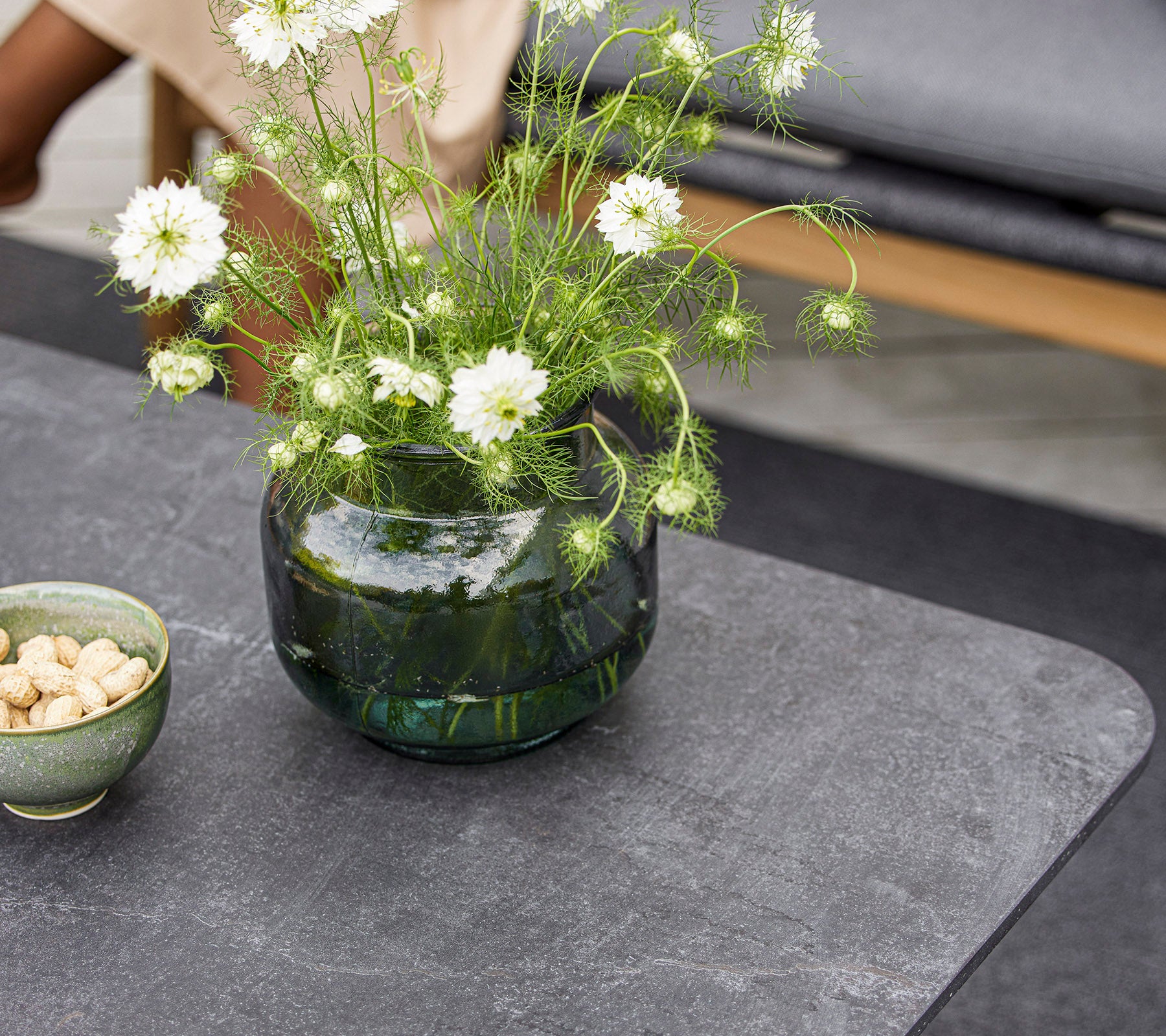 Et moderne bord med en rund glasvase med hvide blomster og en lille skål med nødder. Overfladen er glat og struktureret.