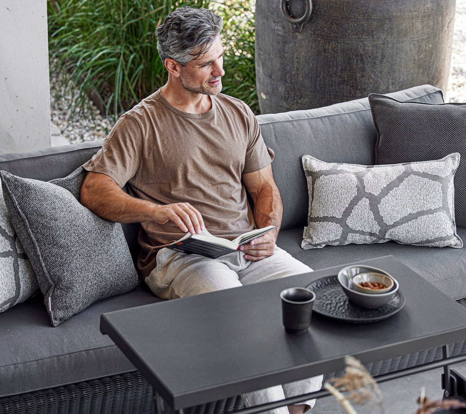 En mand sidder komfortabelt på en sofa, læser en bog med en kop og en skål på et sofabord ved siden af ham, omgivet af dekorative puder.