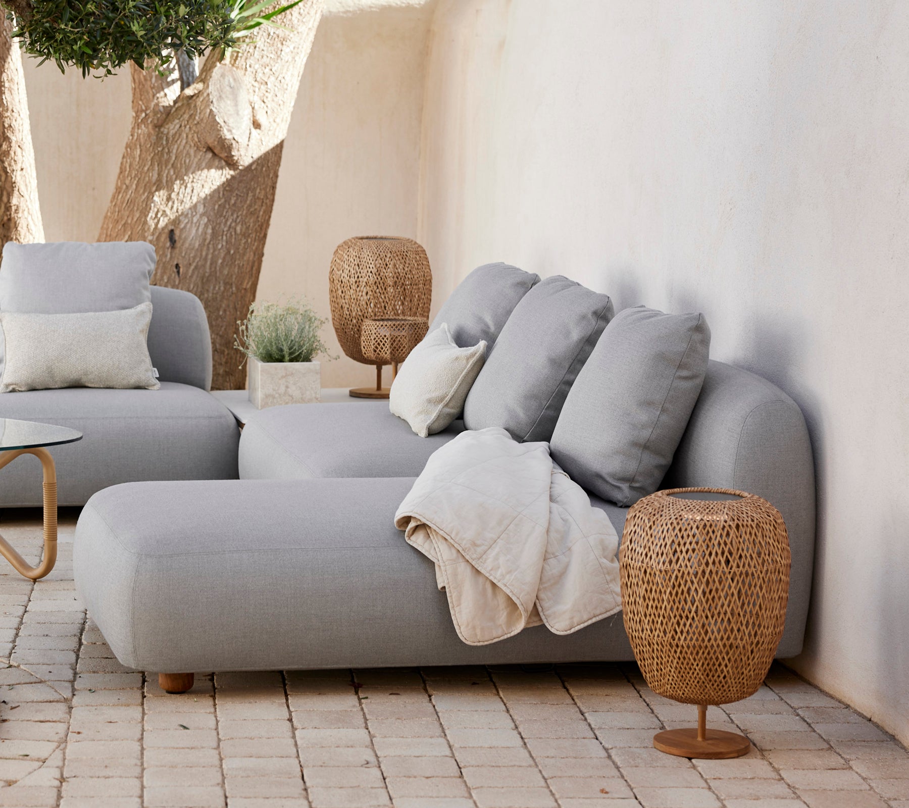 Moderne grå sofa med puder og et vævet sidebord i en minimalistisk stueindretning.