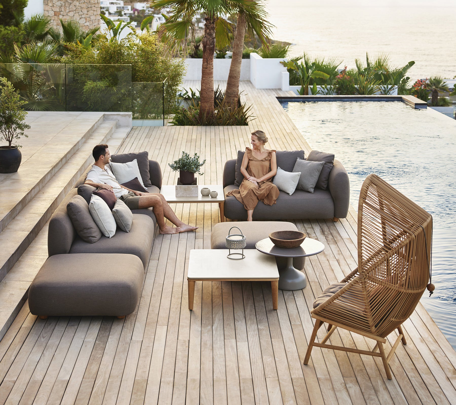 Moderne udendørs loungeområde med grå sofaer, et hvidt bord og en rattanstol.