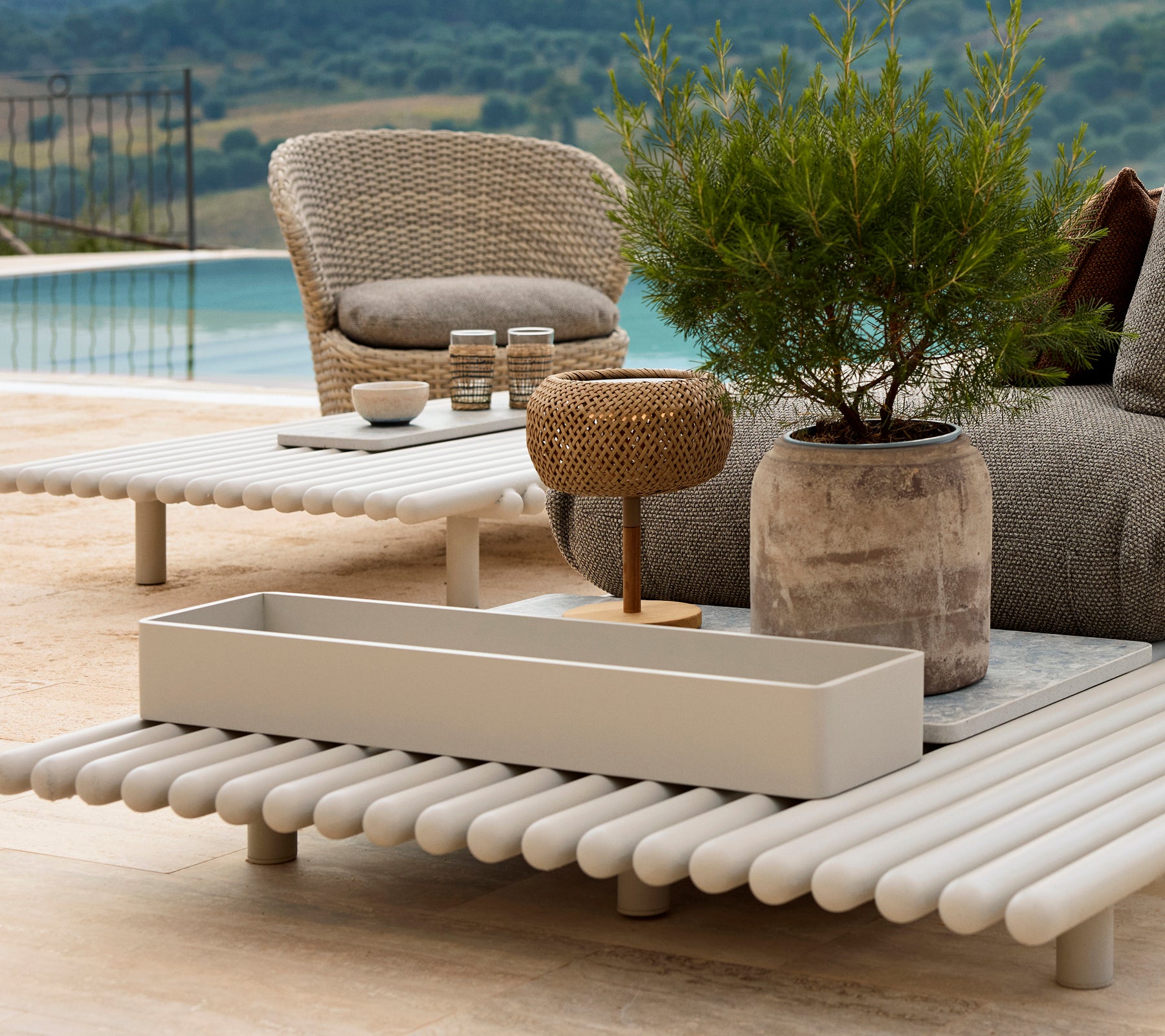 Aluminiums opbevaringsboks på et slatted bord, omgivet af havemøbler og grønne planter, med en naturskøn udsigt i baggrunden.