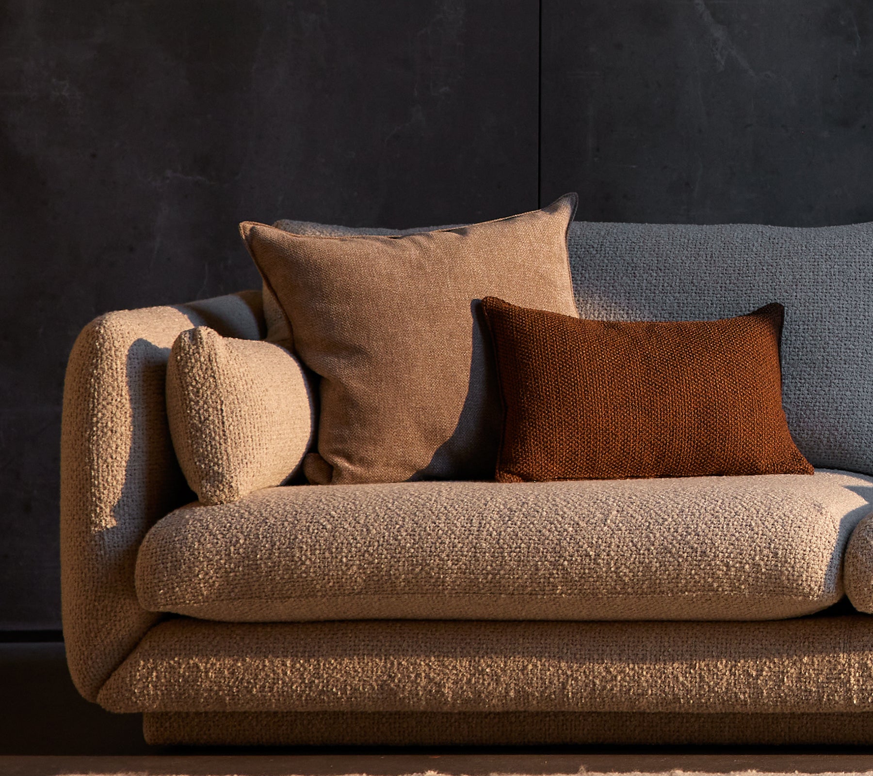 Essence og Rise pyntepuder placeret på en struktureret Bloom-sofa, der har neutrale toner mod en mørk baggrund.