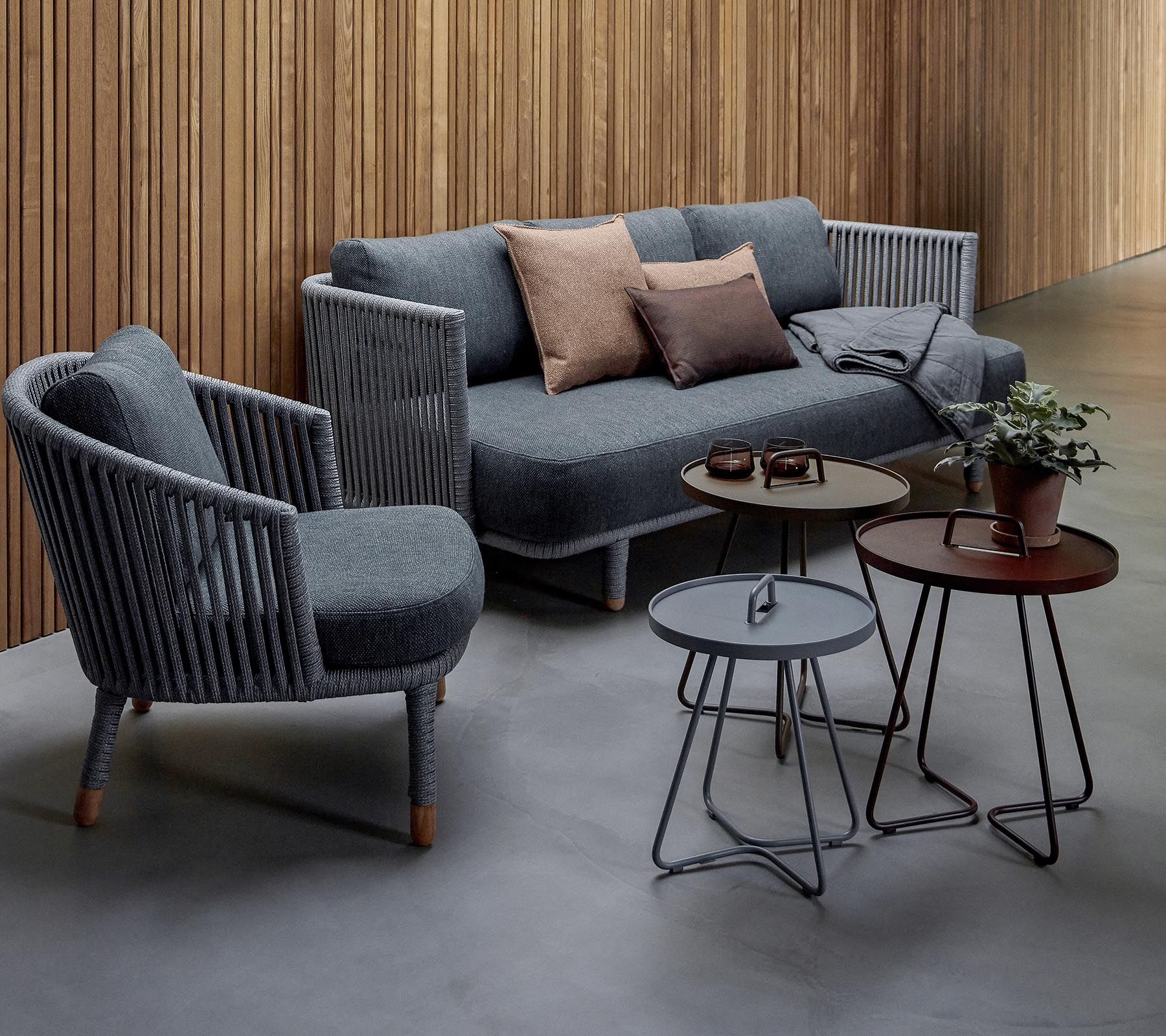 Stilfuld siddearrangement med en grå sofa med puder og to accentstole, ledsaget af tre moderne borde og en potteplante.