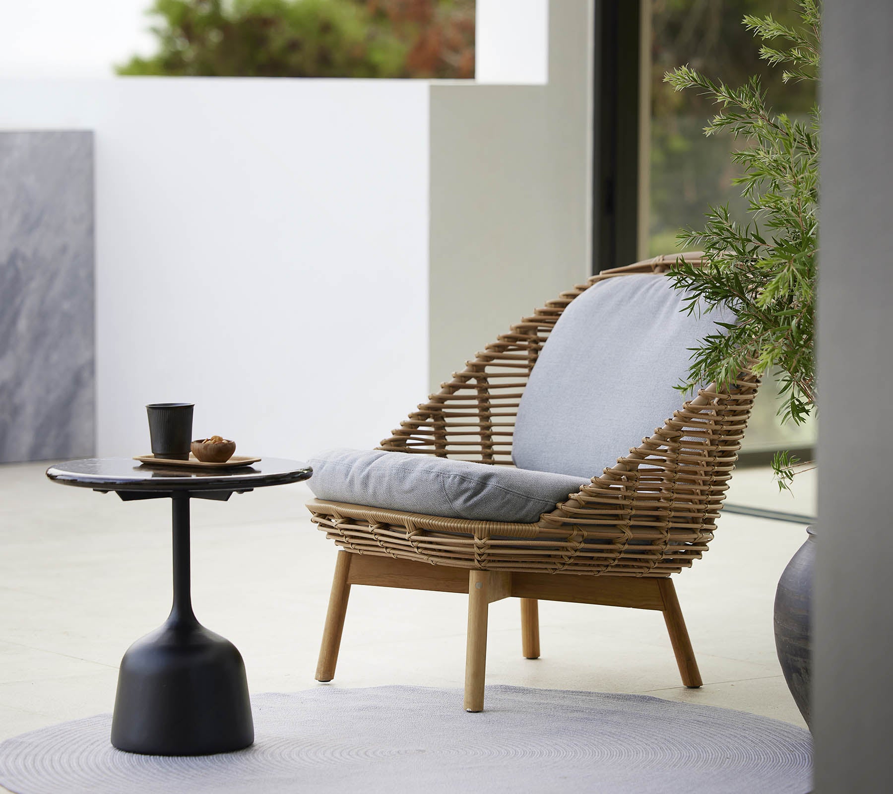 En stilfuld loungestol med et vævet design og bløde puder, ledsaget af et slankt sidebord og en lille plante. Ideel til afslapning.
