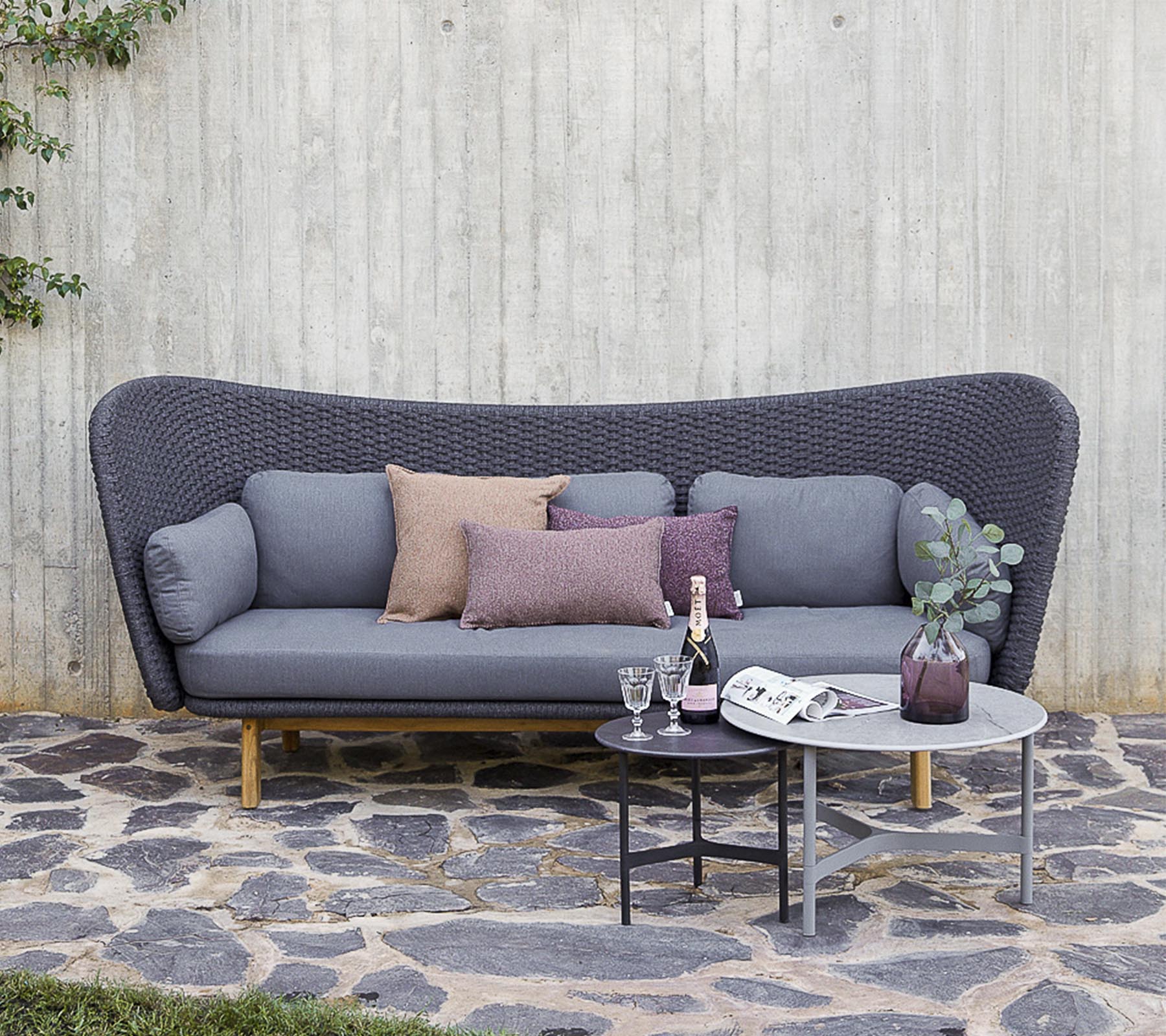 En moderne sofa i grå med dekorative puder, ledsaget af et lille bord med drikkevarer og en vase, sat mod en struktureret væg.