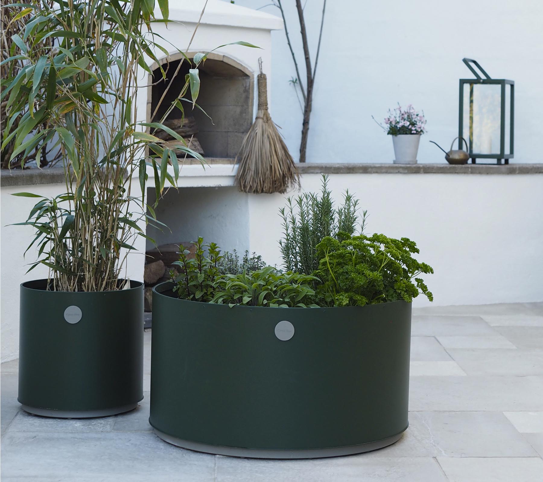 To runde plantekasser i mørkegrøn, den ene med høje planter og den anden fyldt med forskellige urter, står på en lys stensurface.