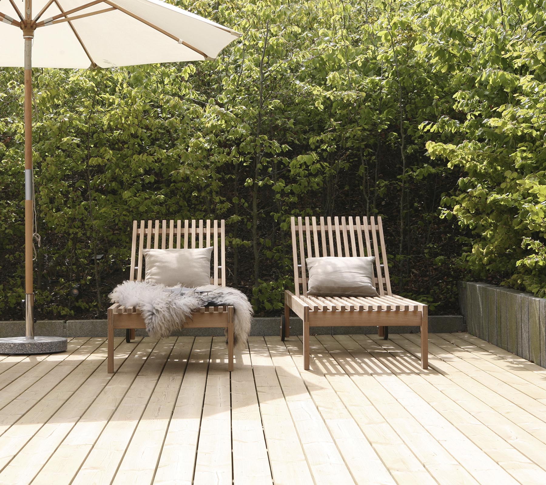 To stilige træloungestole med puder, placeret på en træterrasse, omgivet af grønne planter og en stor parasol i nærheden.