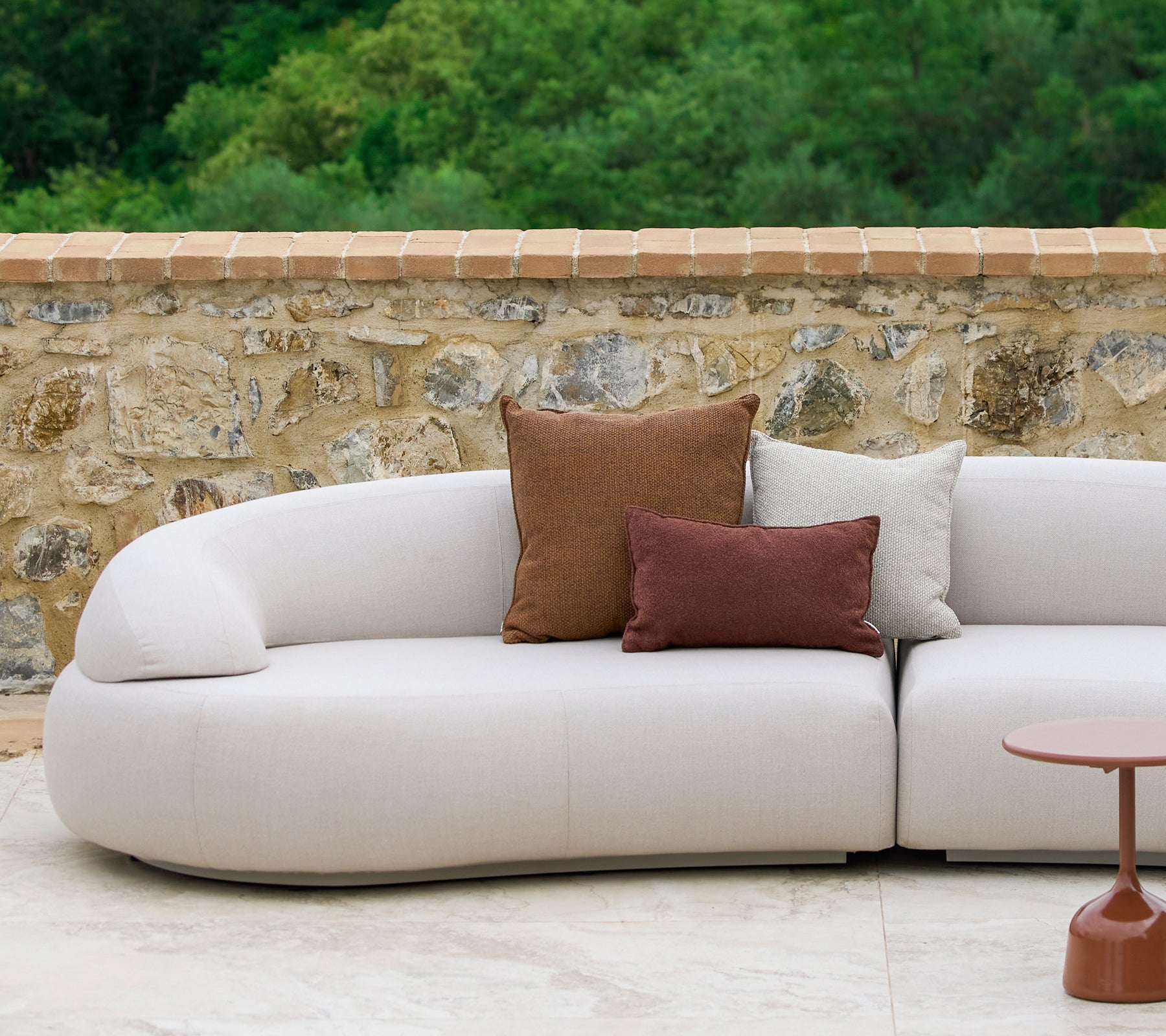 En buet sofa pyntet med Rise puder i jordfarver sammen med et stilfuldt Glaze sidebord, placeret mod en stenbaggrund.