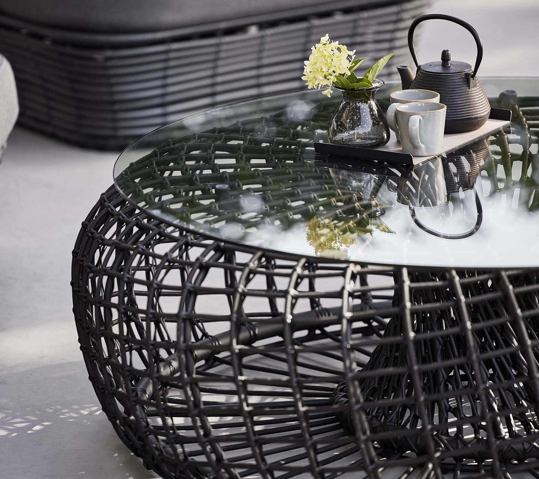 Næstekaffebord med en vævet bund, en glasplade og et tesæt med blomster til et stilfuldt, moderne look.