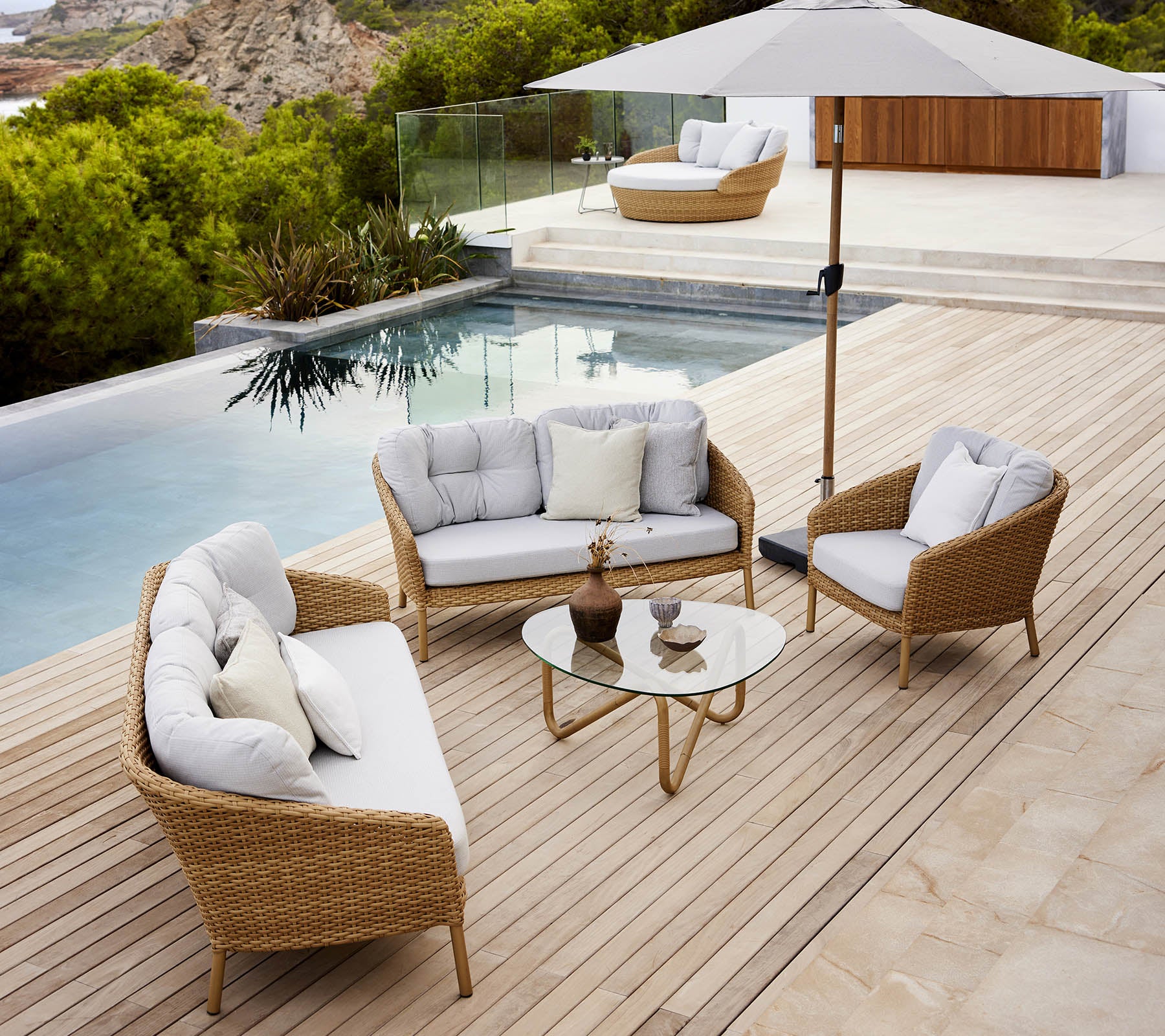 Moderne udendørs loungeområde med et glasbord, komfortable siddepladser og en parasol ved en poolkant med frodig beplantning.