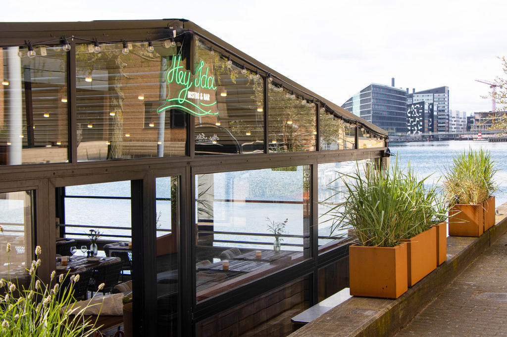 Hey IDA restaurant bygning med grønt neon skilt med Hey IDA, kobber plantekasser med højt græs, udsigt til Kalvebod Brygge i København