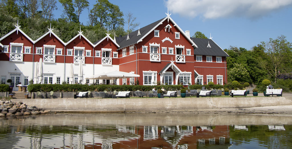 Dyvig Badehotel facade. Rød bygning med hvide detaljer. Udendørs lounge område med udsigt til vandet omgivet af grøn natur og blå himmel